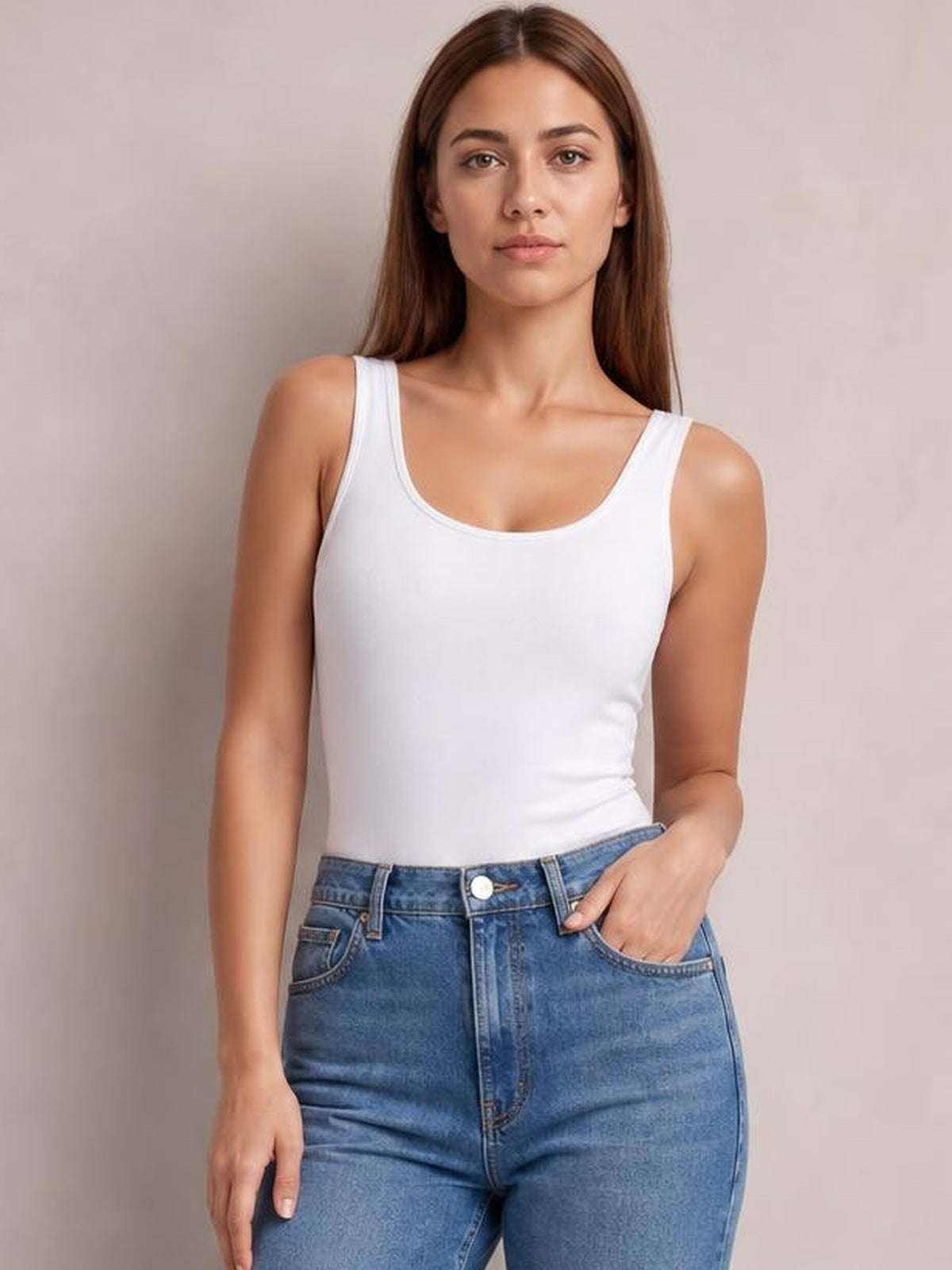 Model wearing a solid white round neck tank with wide shoulder straps and a close-fitting, slightly stretchy silhouette, posing with one hand in her jeans pocket.