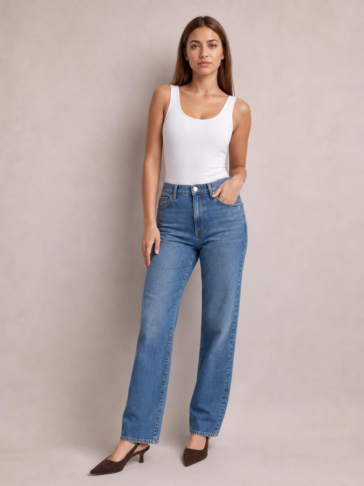 A woman stands in a white solid round neck tank with wide shoulder straps and a fitted silhouette, paired with jeans and one hand in her pocket.