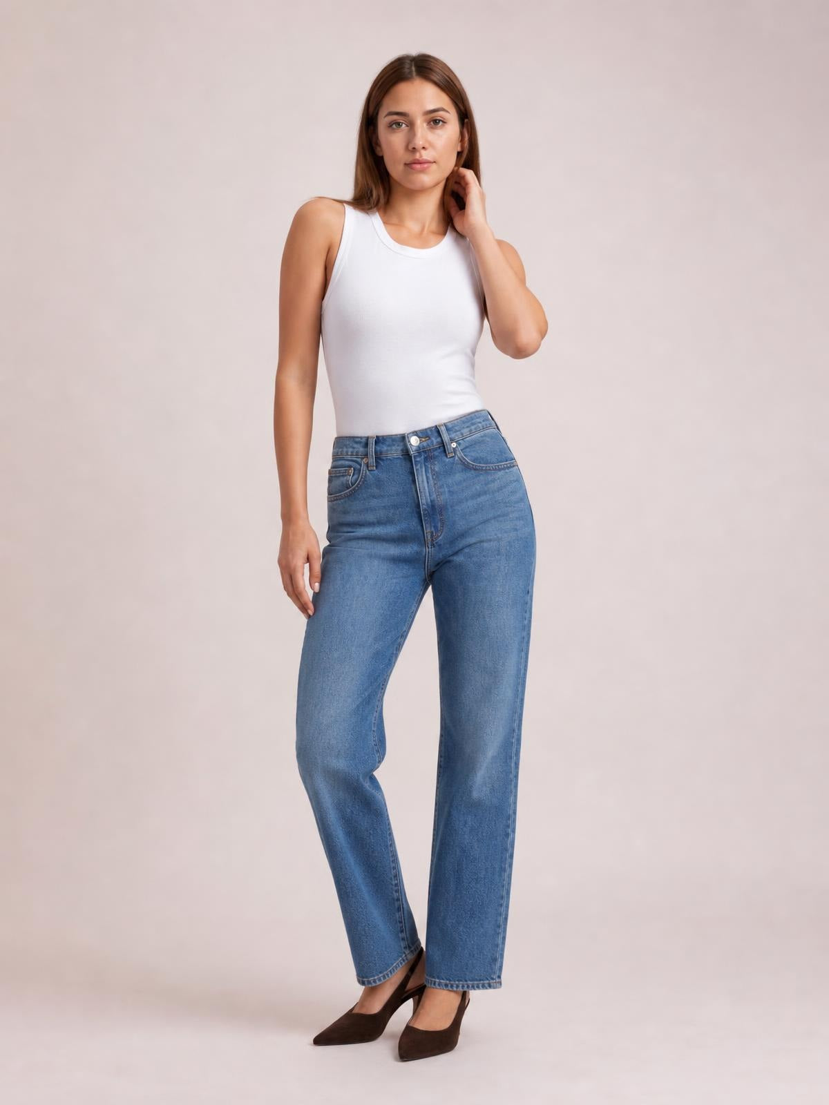 Model wearing a solid white round neck tank with a fitted silhouette and wide shoulder straps, posing with one hand touching her hair.