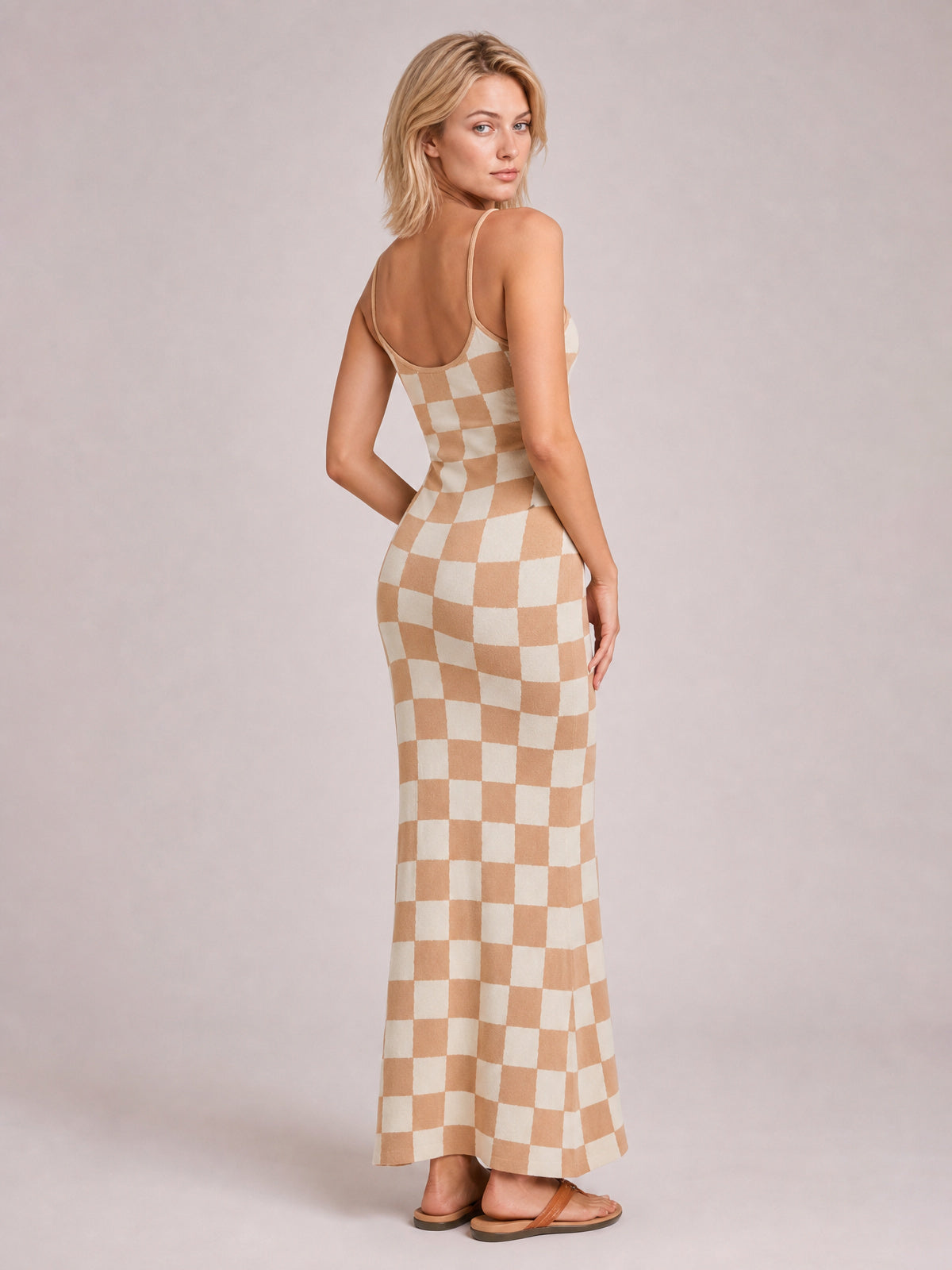 A model looks over her shoulder in a tan and cream checkerboard spaghetti-strap maxi sweater dress with a fitted silhouette and gently flared ankle-length hem.