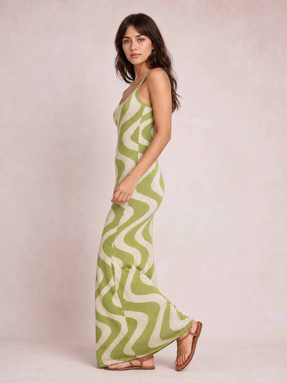 Model in a green and cream patterned spaghetti strap maxi sweater dress with a fitted silhouette, ankle-skimming hem, and side pose in flat sandals.