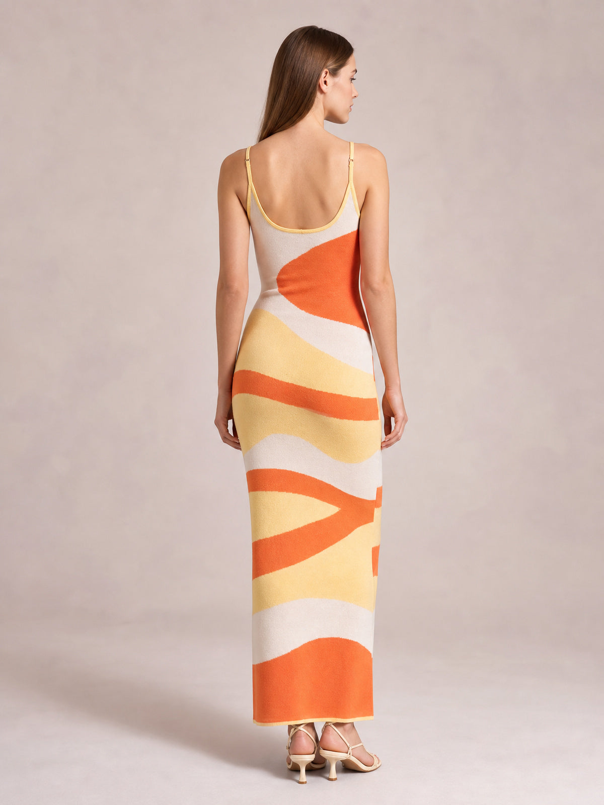 Model stands with her back to the camera in a fitted spaghetti strap maxi sweater dress with orange, yellow, and cream abstract stripes and an ankle-skimming hem.