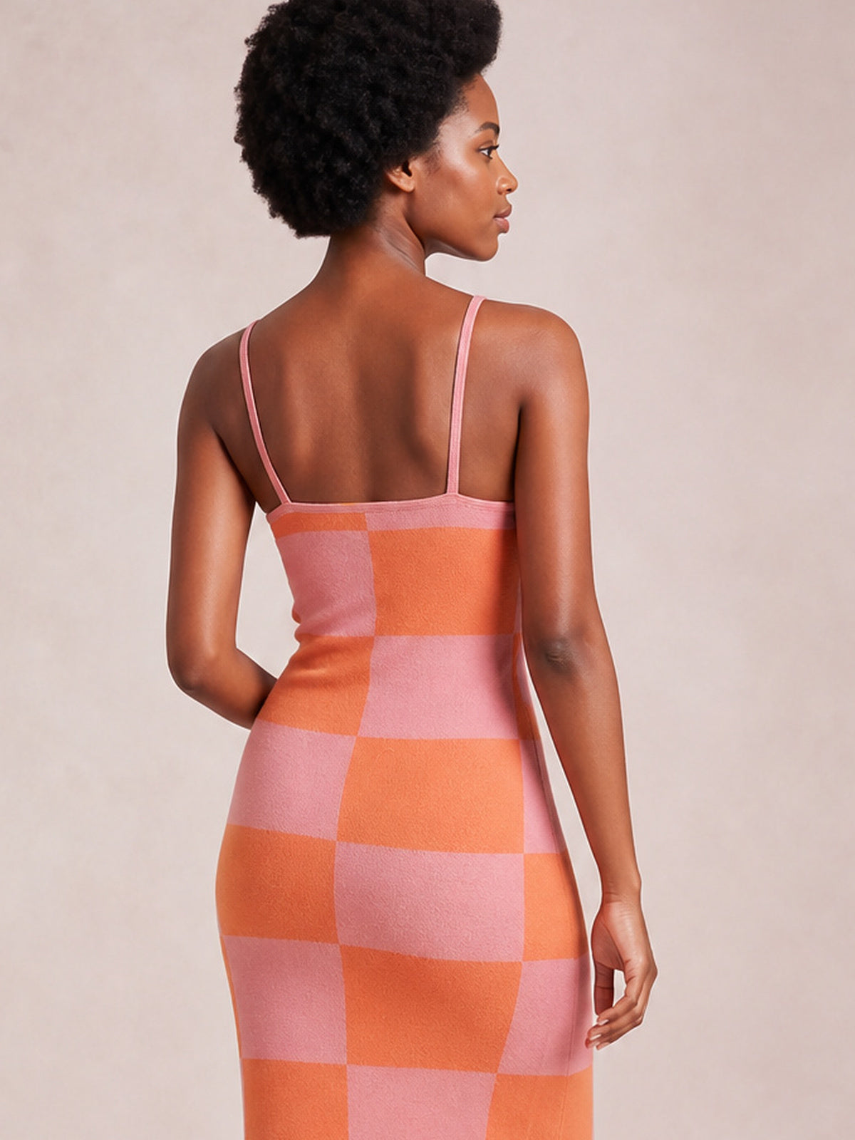 A model looks over her shoulder in a fitted pink and orange checkerboard spaghetti strap maxi sweater dress with a square back neckline and long silhouette.