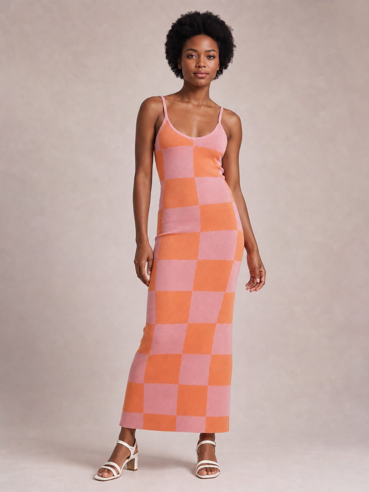 A model stands in a fitted pink and orange checkerboard spaghetti strap maxi sweater dress with an open neckline and ankle-skimming hem.