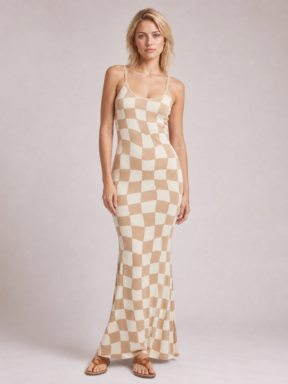 A model stands in a beige and cream checkerboard maxi sweater dress with spaghetti straps, scoop neckline, fitted silhouette, and gently flared ankle-length hem.