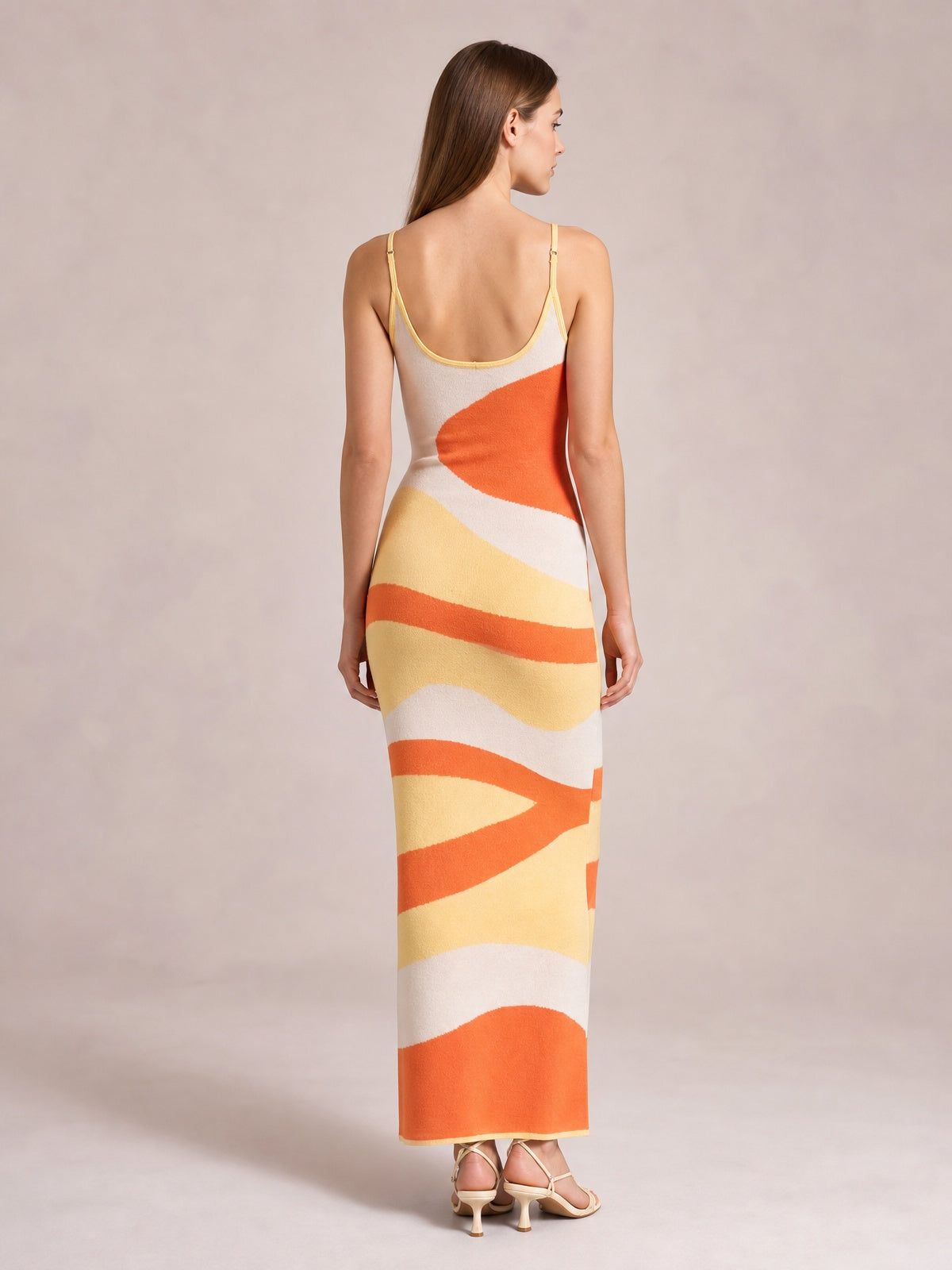Model stands with her back to the camera in a fitted spaghetti strap maxi sweater dress with orange, yellow, and cream abstract stripes and an ankle-skimming hem.