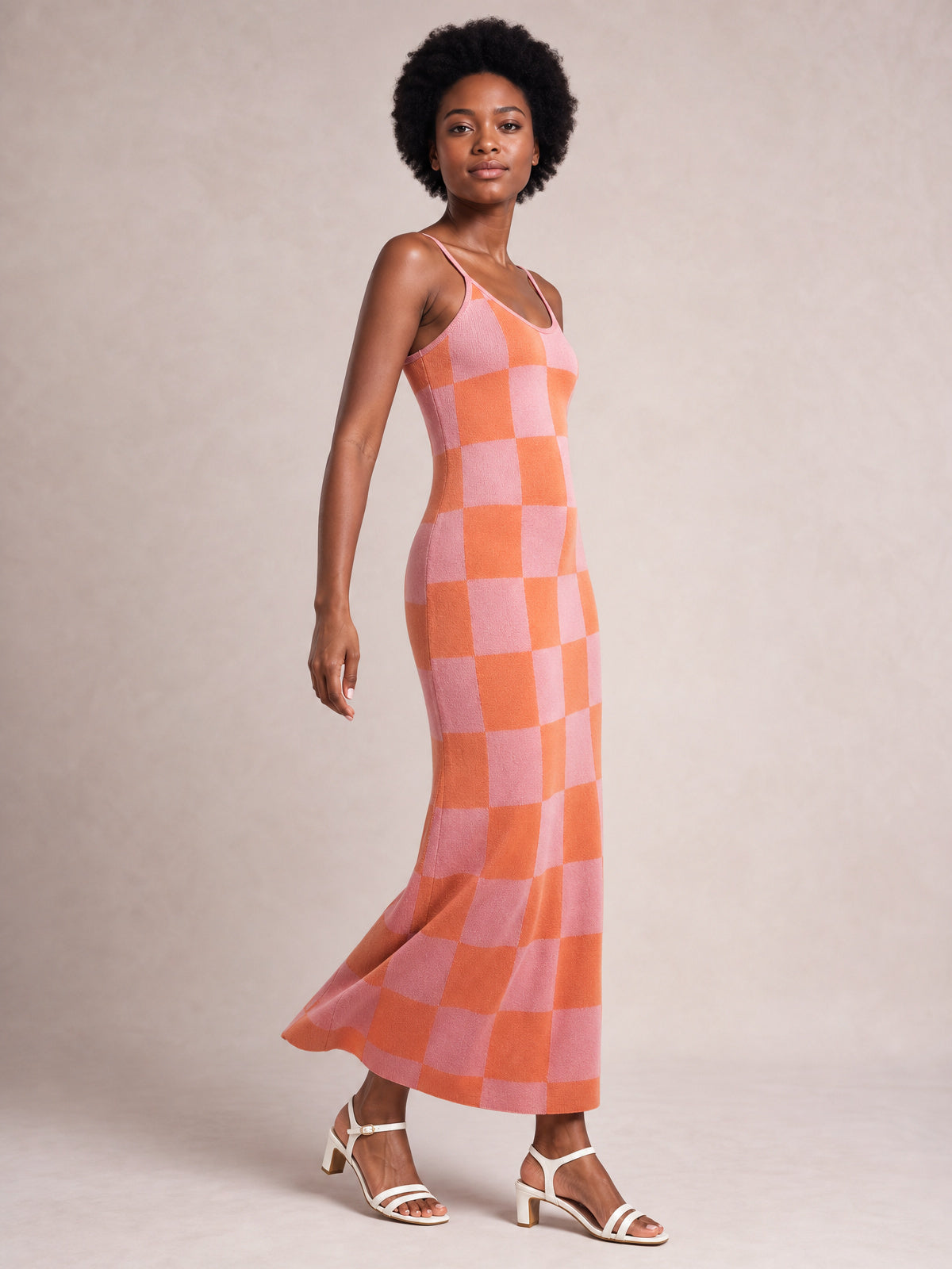 A model poses in a pink and orange checkerboard spaghetti strap maxi sweater dress with a fitted silhouette and gently flared ankle-skimming hem.