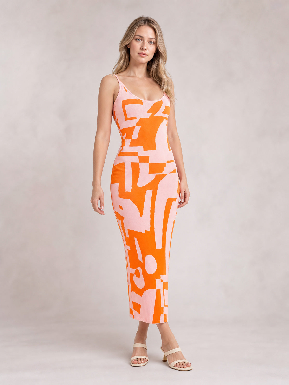 Model poses in a pink spaghetti strap maxi dress with bold orange abstract print, a fitted silhouette, open neckline, and ankle-skimming hem.