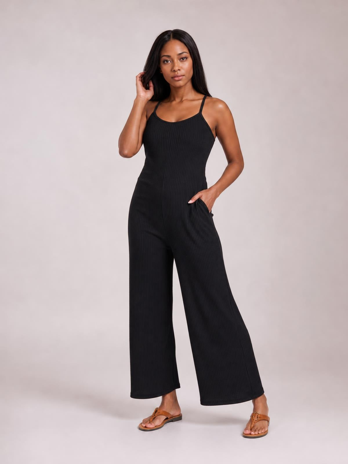 Model poses in a black spaghetti strap V-neck jumpsuit with a fitted bodice, wide-leg silhouette, vertical ribbed texture, and side pockets.