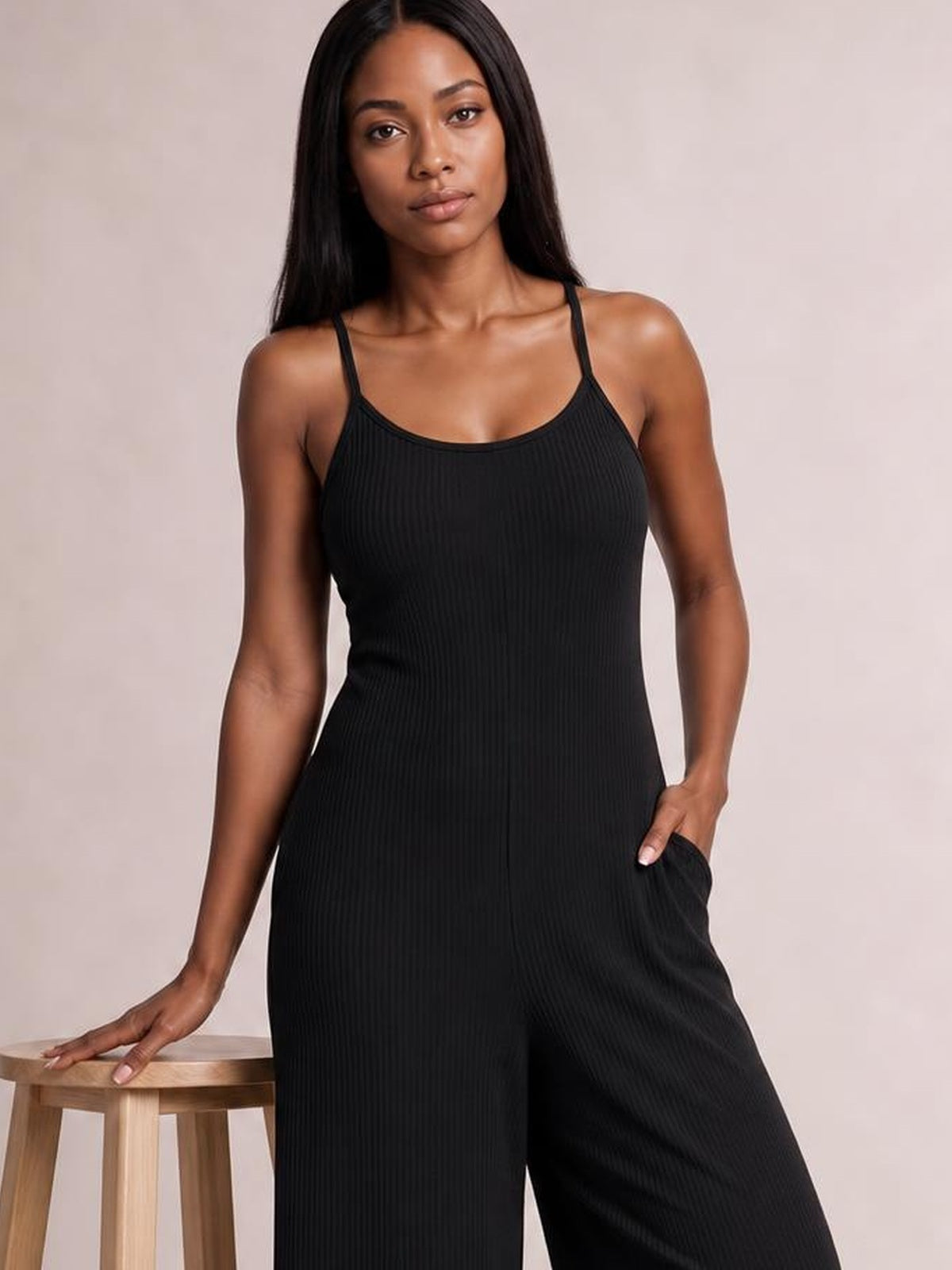 Model wears a black spaghetti-strap jumpsuit with a fitted ribbed bodice, wide-leg silhouette, and a visible side pocket, posing with one hand resting on a stool.