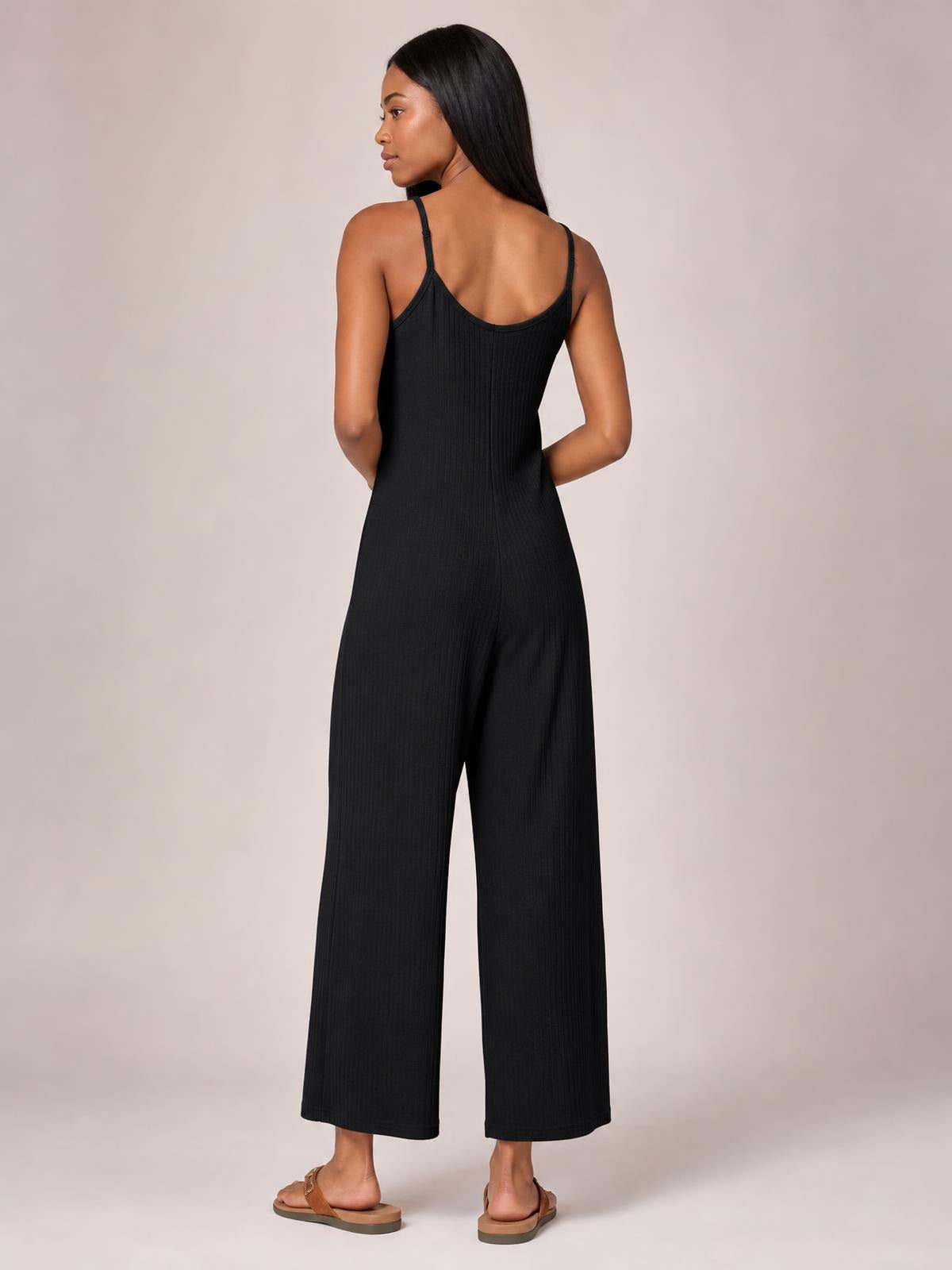Model shown from behind in a black spaghetti strap jumpsuit with a fitted bodice, scoop back, subtle ribbed texture, and wide-leg silhouette.