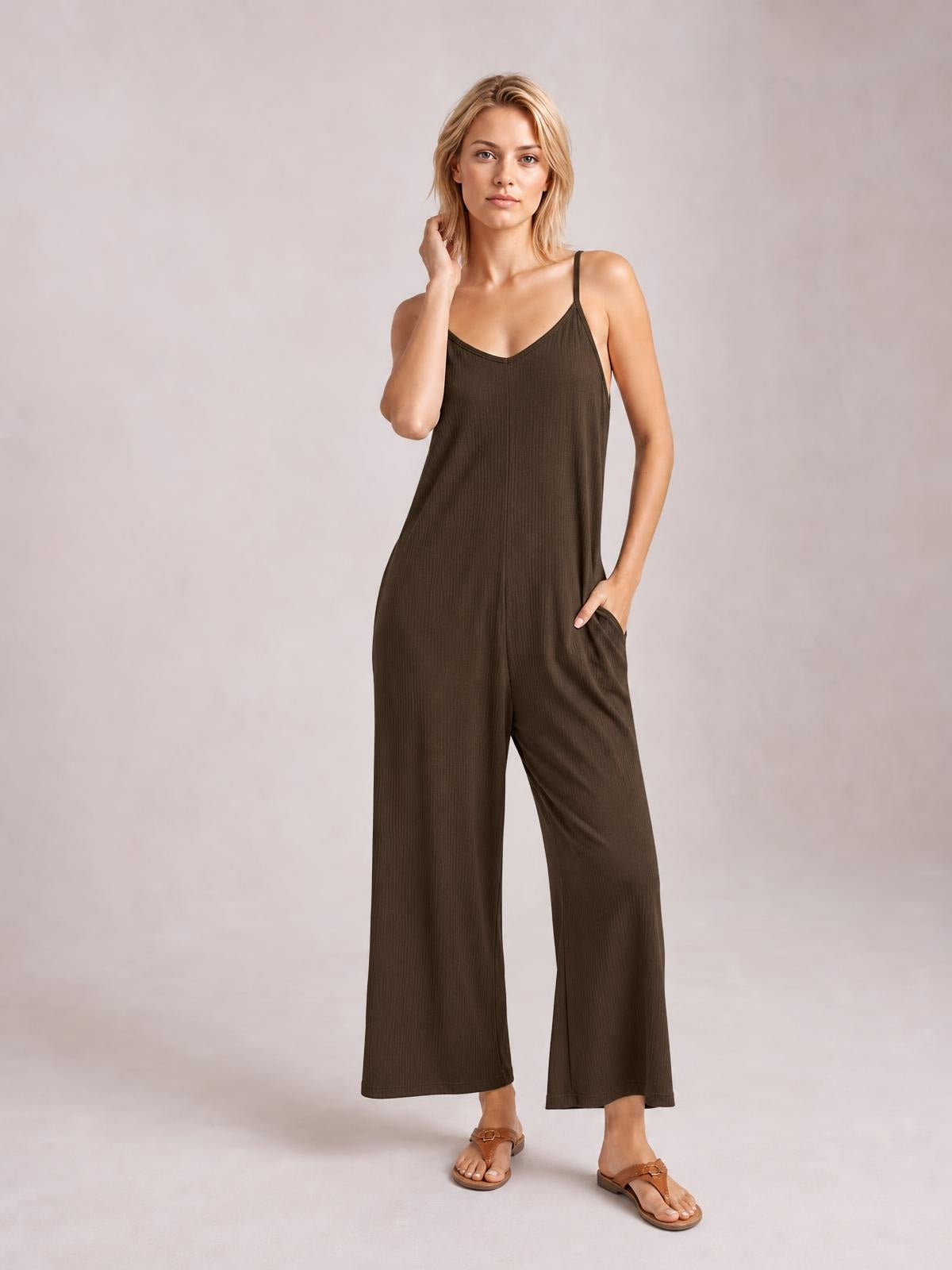 Model poses in a brown spaghetti strap V-neck jumpsuit with a relaxed wide-leg silhouette and side pockets, one hand resting at her neck and the other in a pocket.