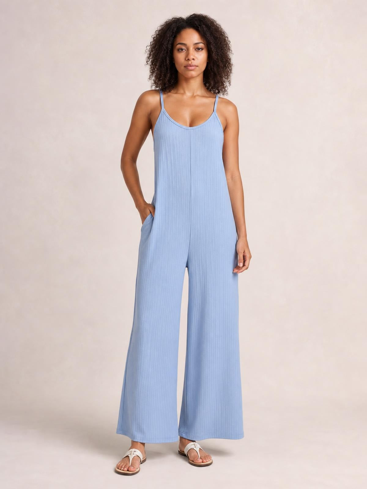 A model wears a light blue spaghetti-strap V-neck jumpsuit with a ribbed texture, relaxed wide-leg silhouette, and side pockets, posing with one hand tucked in.