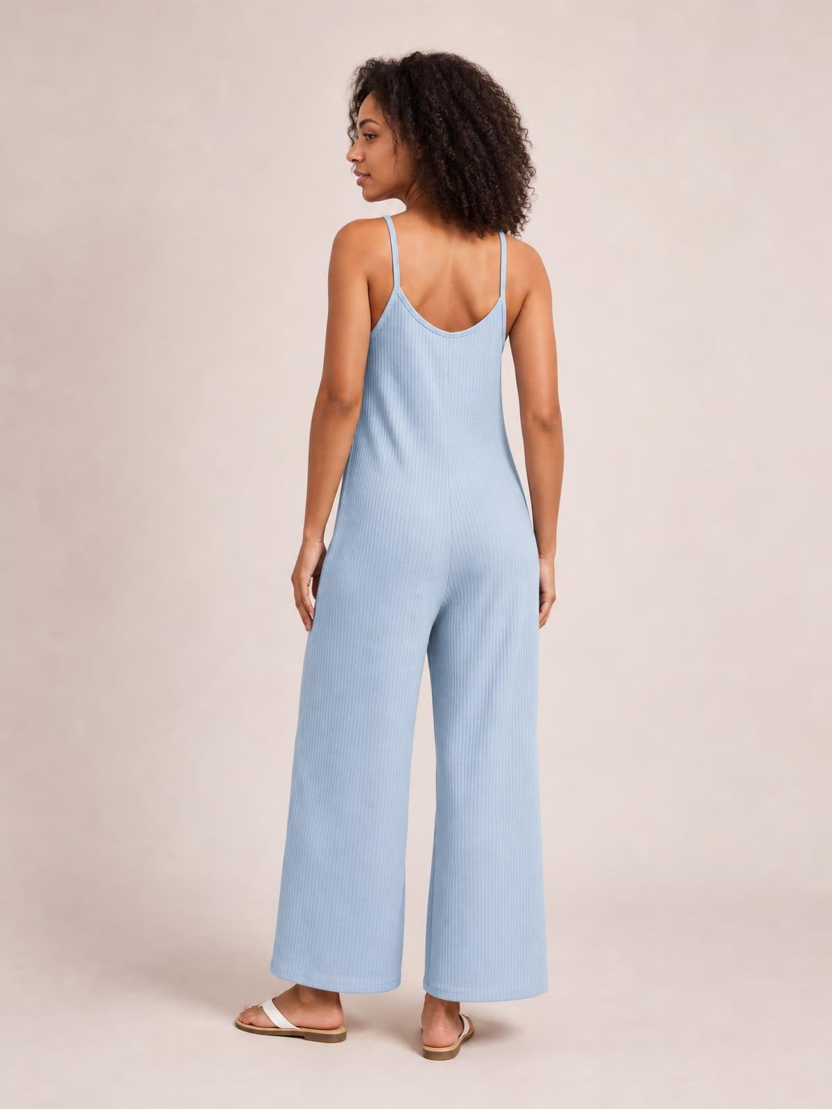 Model shown from the back in a light blue spaghetti strap jumpsuit with a low scoop back, ribbed texture, fitted waist, and wide-leg silhouette.