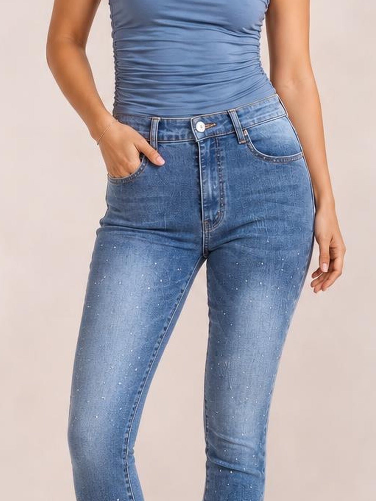 A model poses with one hand in the pocket, wearing blue high-waisted skinny jeans with scattered rhinestones, front pockets, belt loops, and a zip fly.