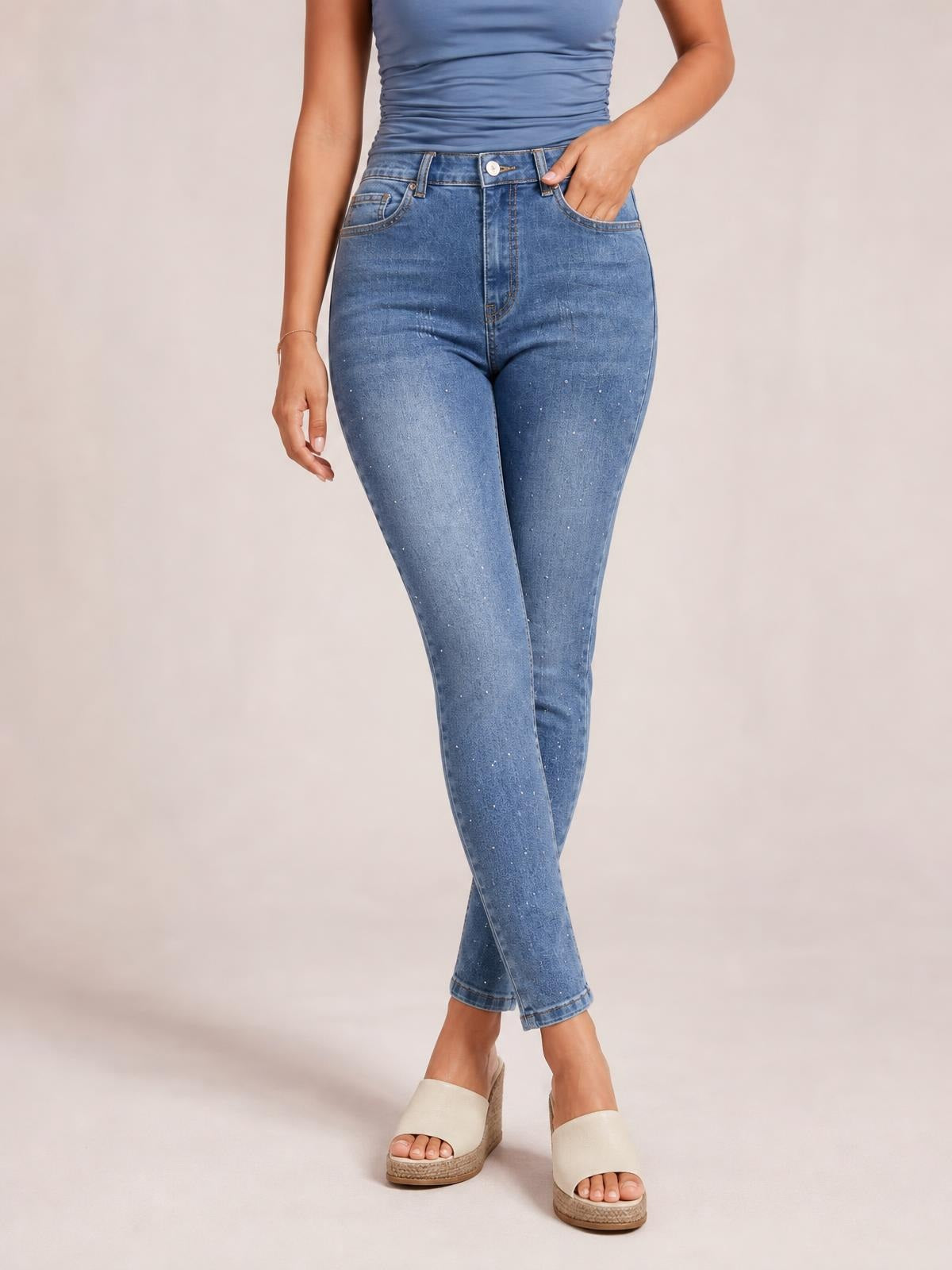 Model poses in sparkling rhinestone high-waist skinny jeans with a blue denim wash, zip fly, belt loops, front pockets, and a close-fitting tapered silhouette.
