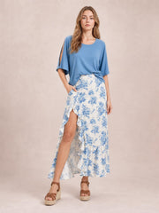 Model wearing a white and blue floral printed skirt with an asymmetrical split, layered ruffled trim, and flowing midi-length silhouette.