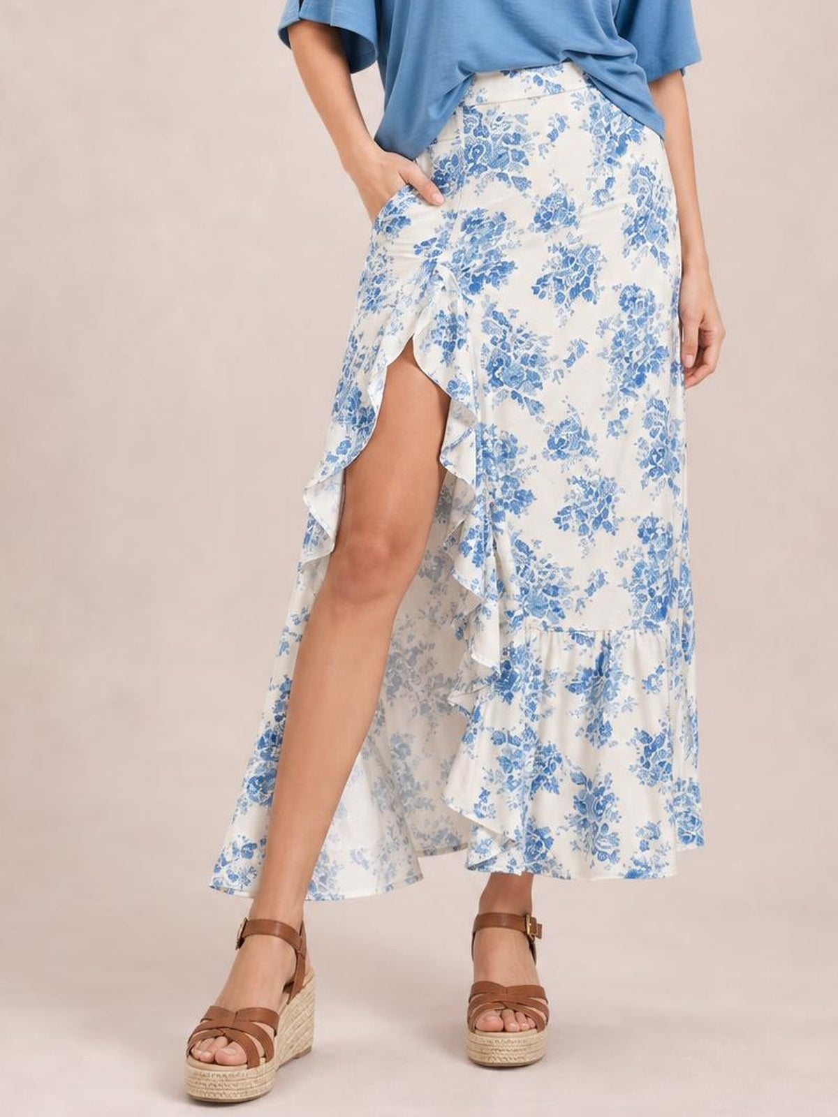 A model poses in a white and blue floral printed skirt with an asymmetrical split, cascading ruffles, and a flowing ankle-length silhouette.