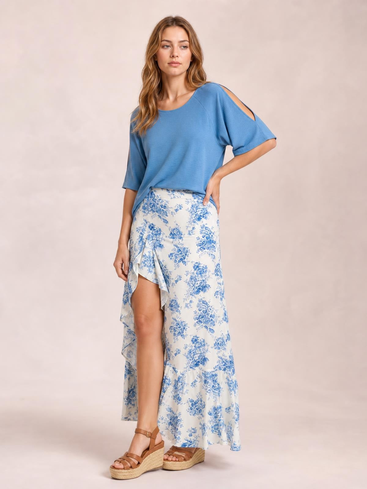 A model poses in a white and blue floral printed skirt with an asymmetrical split, layered ruffled hem, and flowing maxi silhouette.