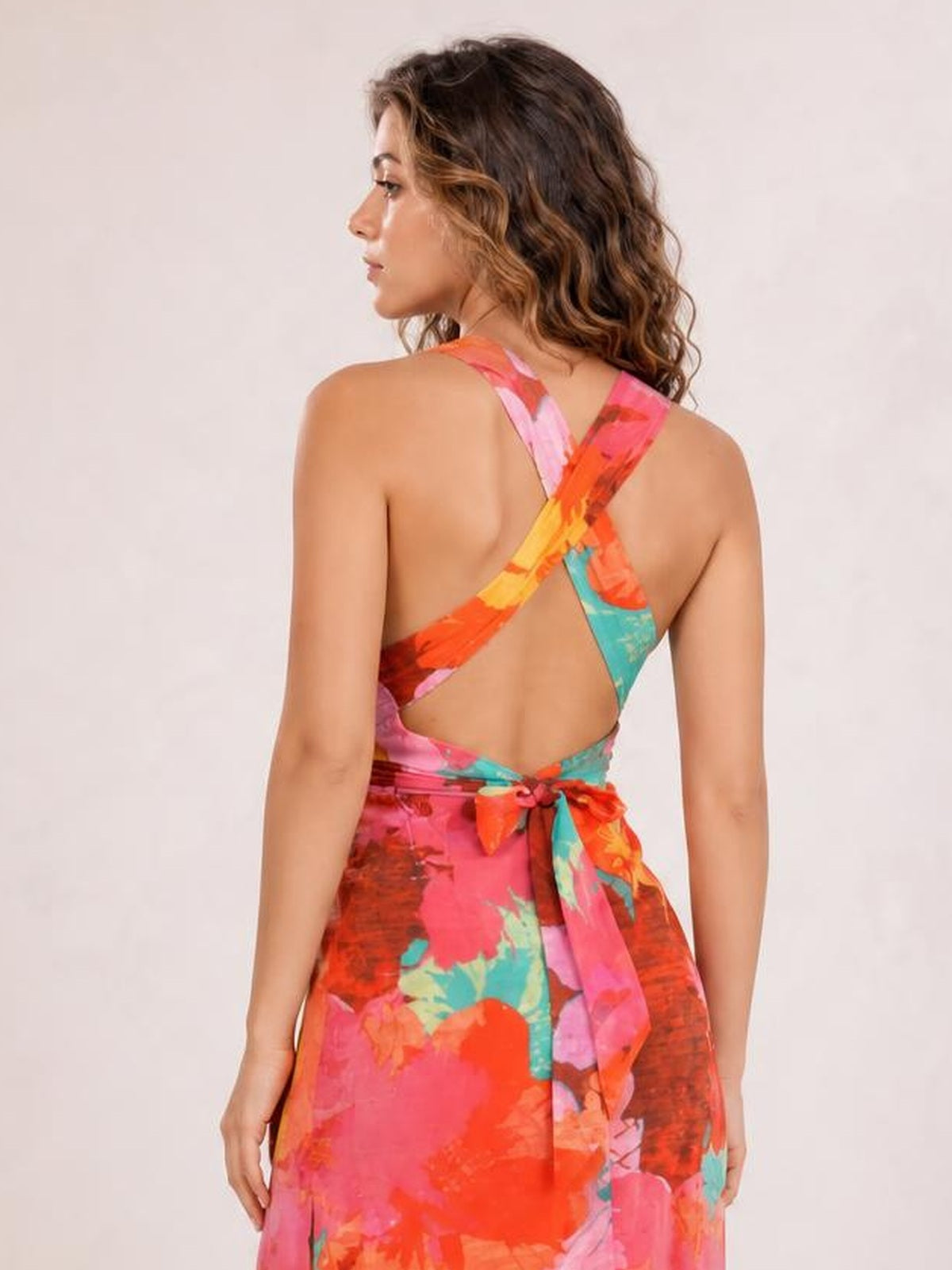 A model poses in a vibrant printed maxi dress with a crisscross open back, tied waist sash, and flowing silhouette in pink, orange, turquoise, and red.