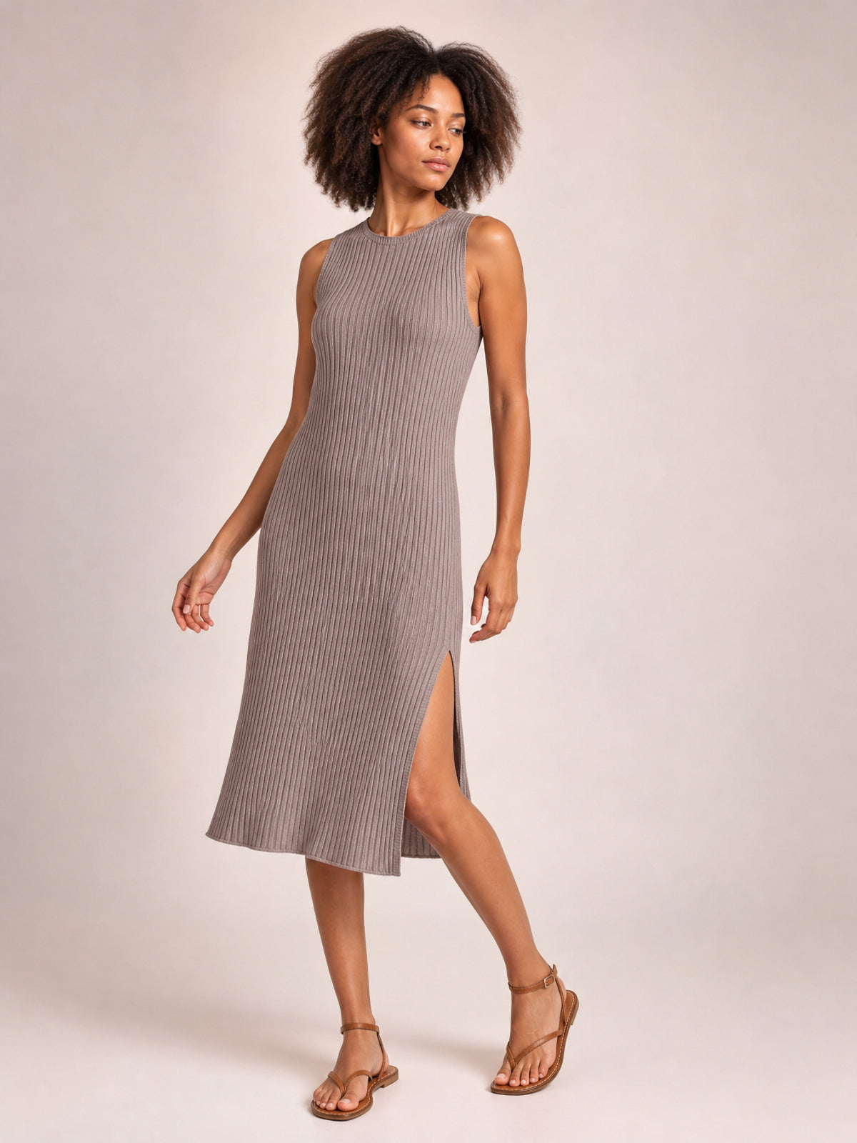 Model poses in a taupe sleeveless midi dress with a round neck, vertical ribbed texture, body-skimming silhouette, and side split.