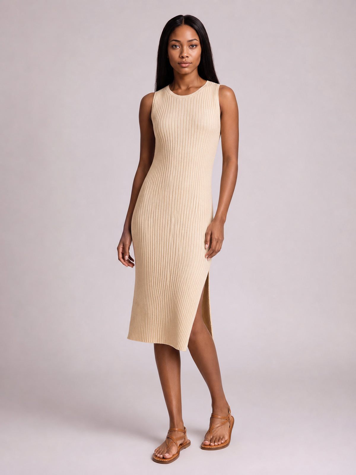 Model poses in a beige sleeveless midi dress with a round neck, body-skimming silhouette, vertical ribbed texture, and side split, paired with flat strappy sandals.