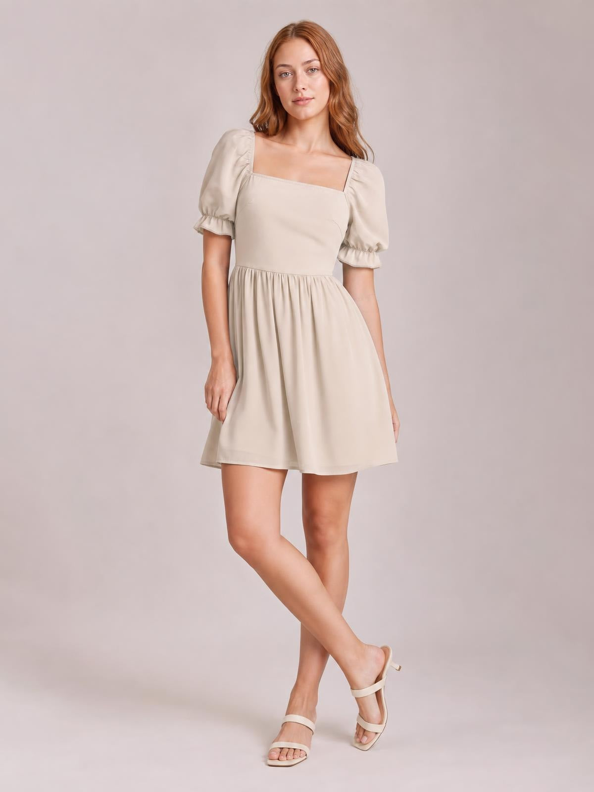 A model poses with crossed legs in a cream square neck mini dress featuring puffed flounce sleeves, a fitted bodice, and a softly gathered skirt.
