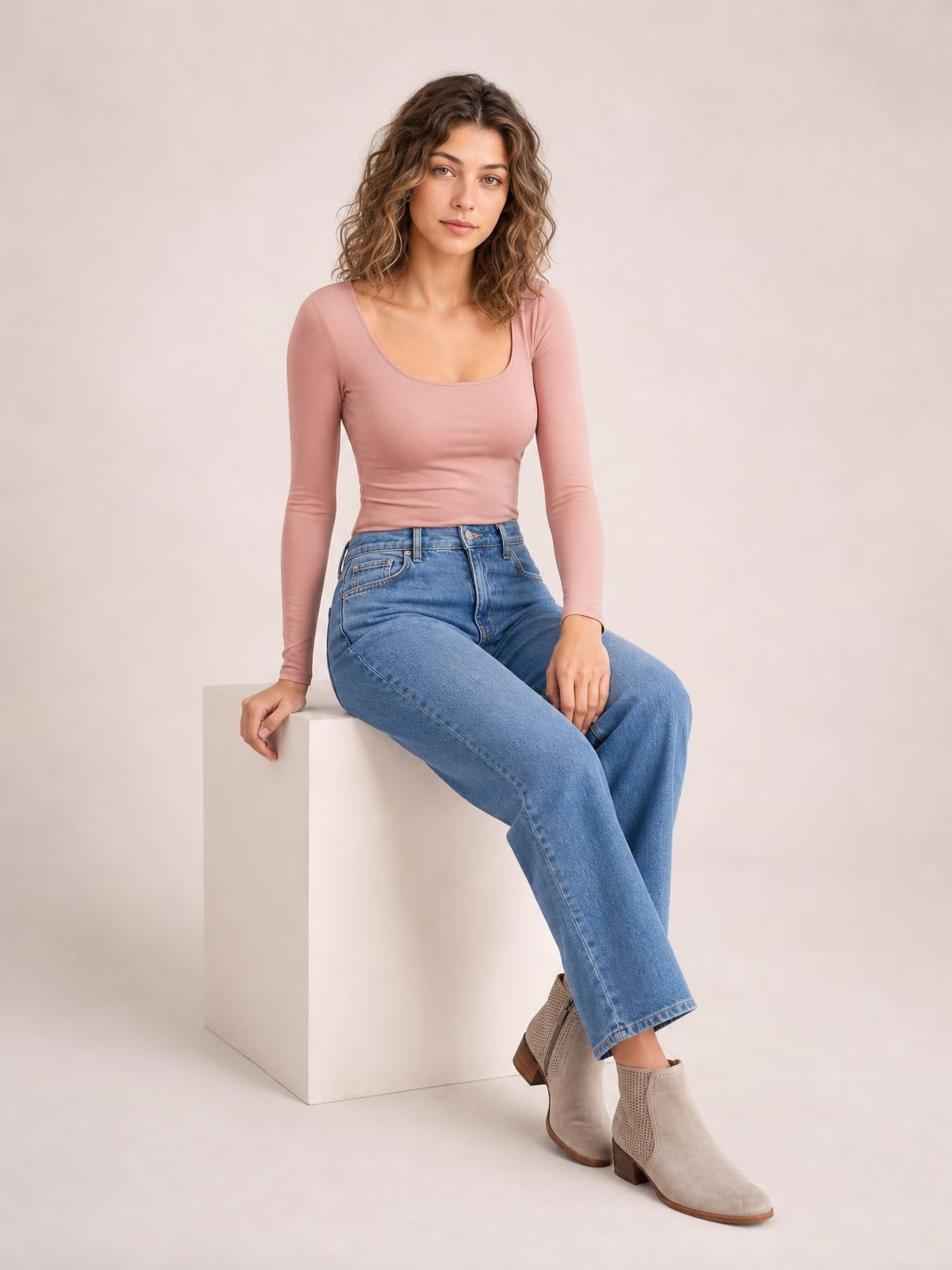 A model sits with one hand resting beside her, wearing a fitted dusty pink square neck long sleeve T-shirt with a smooth, simple silhouette.
