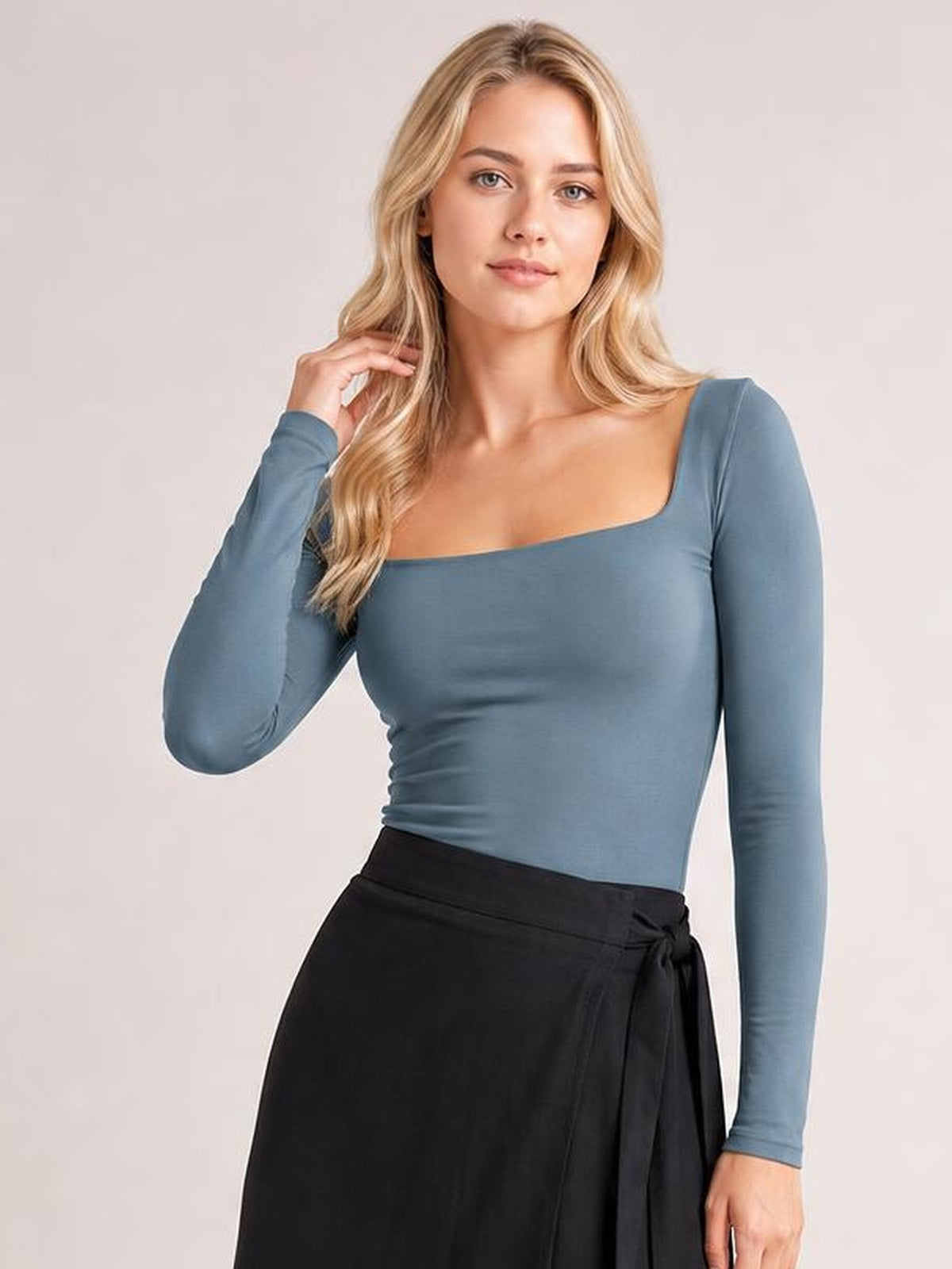 Model wears a blue square neck long sleeve T-shirt with a fitted silhouette and stretch construction, posing with one hand touching her hair.