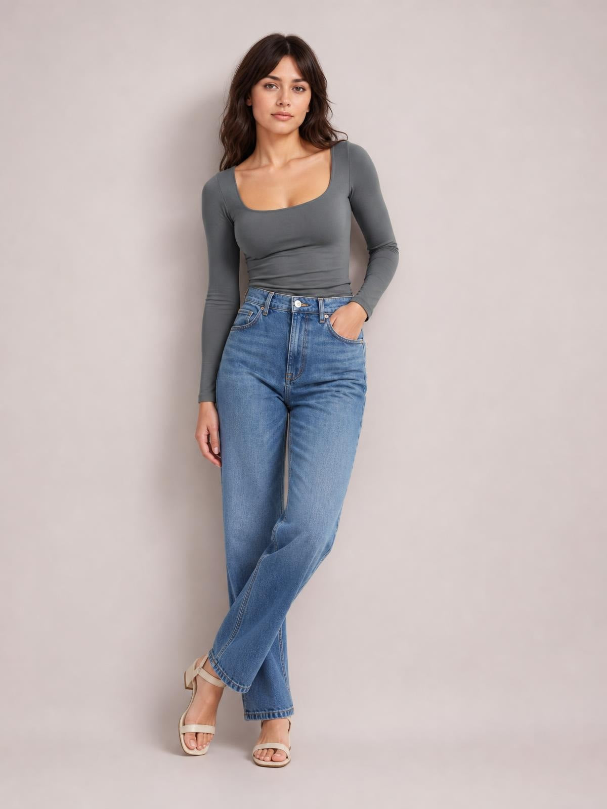 Model wearing a fitted gray square neck long sleeve T-shirt with a smooth, opaque silhouette, paired with jeans while standing with one hand in a pocket.