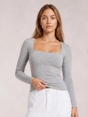 A woman models a gray square neck long sleeve T-shirt with a fitted silhouette, gently lifting the hem to show its stretch and clean construction.