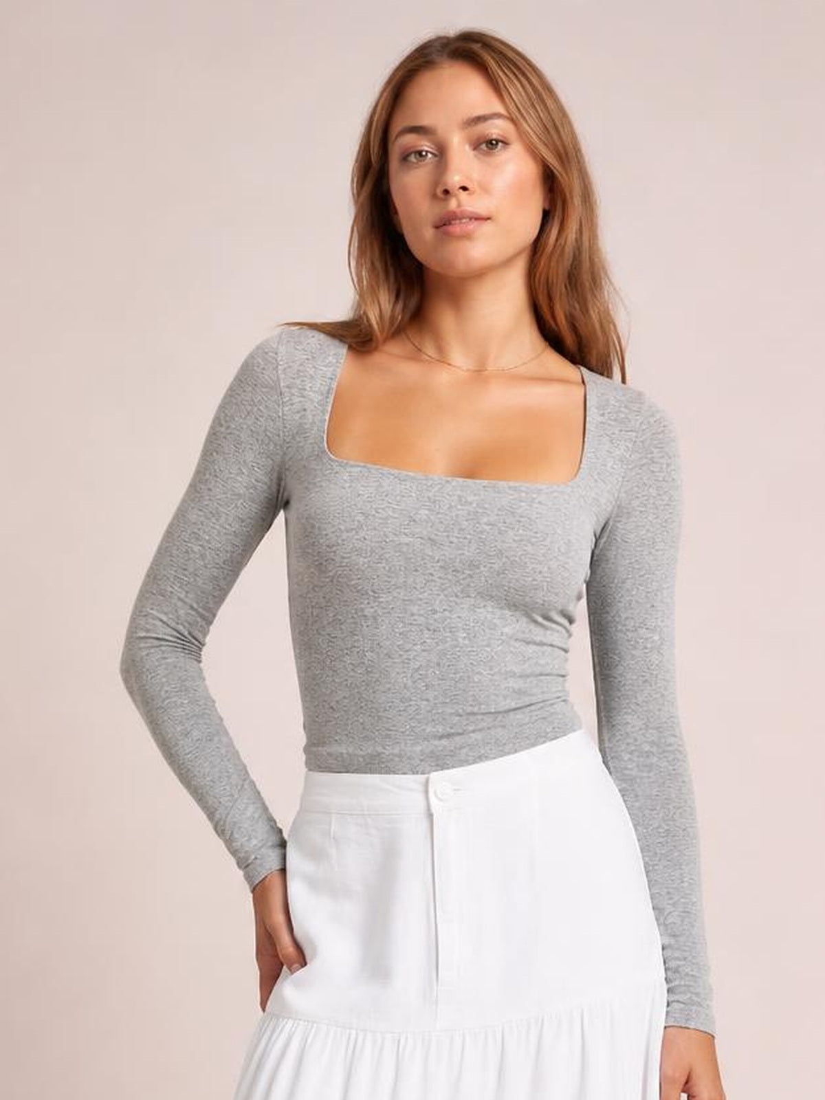 Model wears a gray square neck long sleeve T-shirt with a fitted silhouette, smooth construction, and one hand resting in a white skirt pocket.