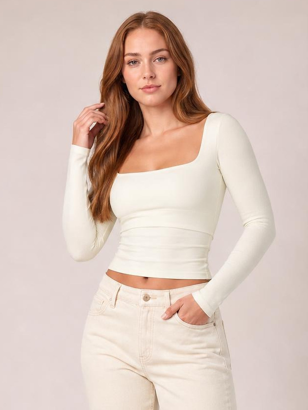 A model poses in a fitted ivory square neck long sleeve T-shirt with a waist-length silhouette, one hand touching her hair and the other in her pocket.