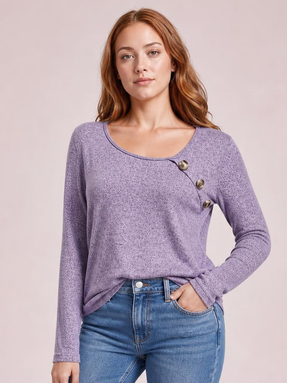 Model wears a lavender long sleeve T-shirt with a rounded neckline, tonal textured pattern, relaxed silhouette, and diagonal button detail, posing with one hand in her jeans pocket.