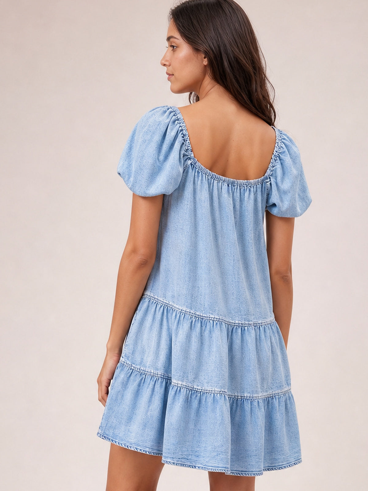 A model turns to show the light denim dress’s ruched square back neckline, puff sleeves, and relaxed A-line silhouette with gathered tier seams.