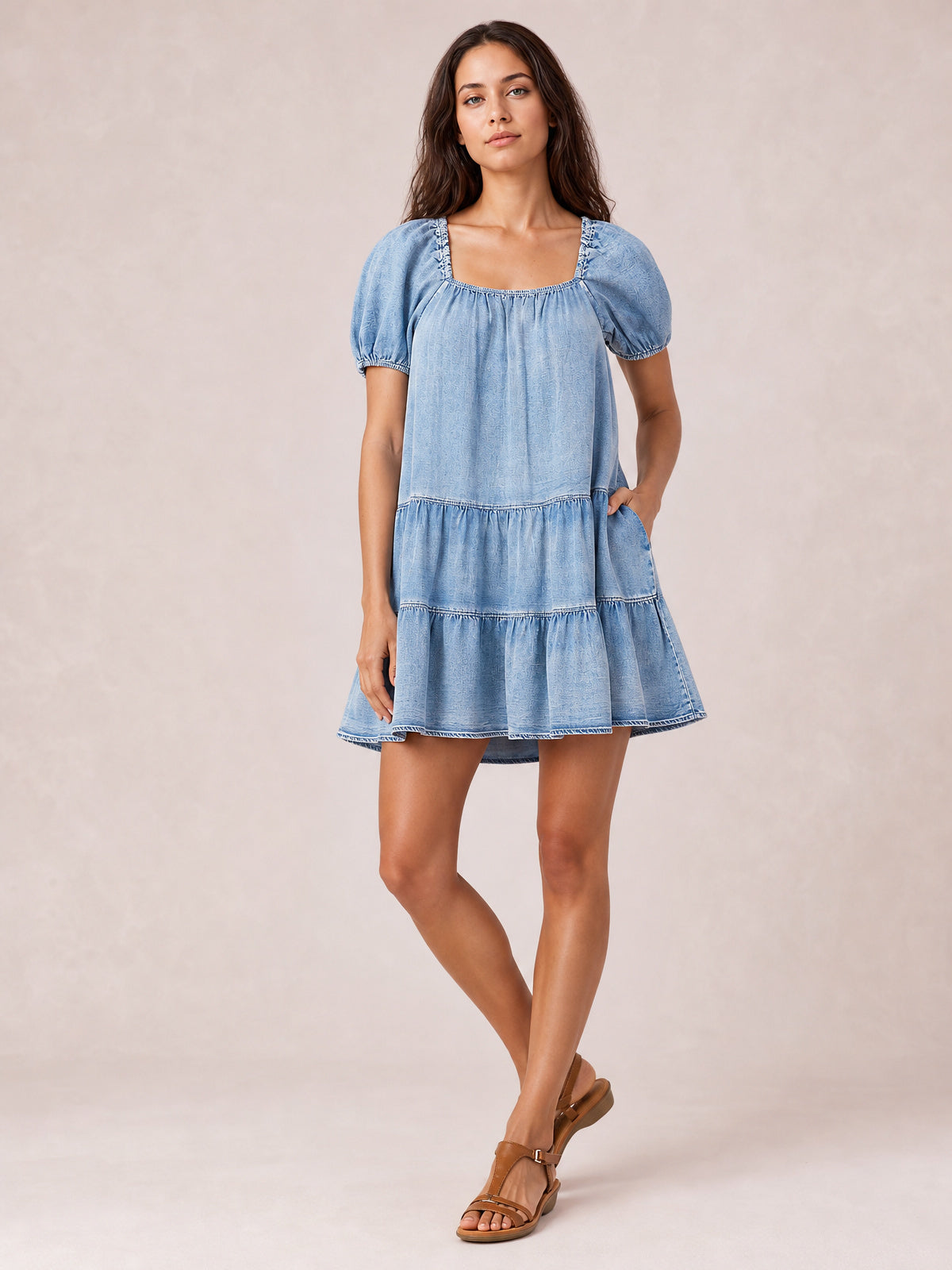 Model poses in a light wash denim dress with a square neck, puff sleeves, gathered tier seams, ruched neckline, and relaxed A-line short silhouette.