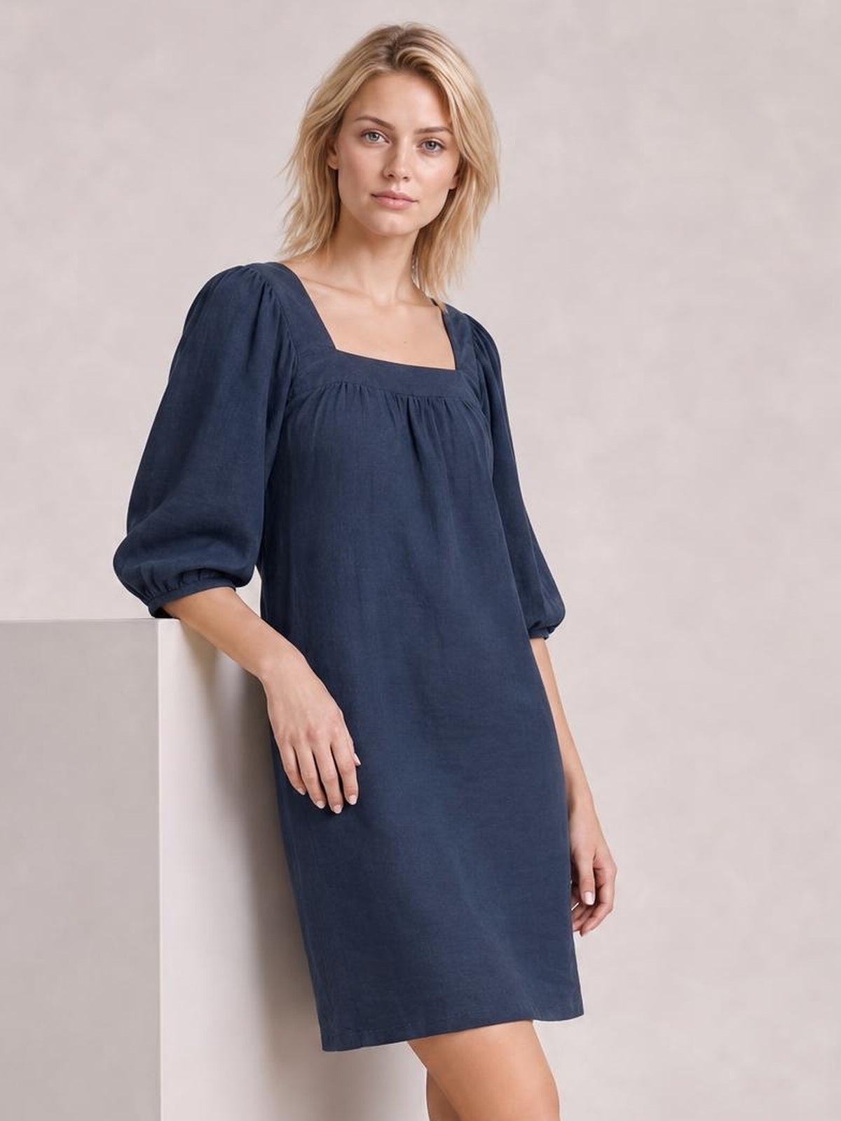 Model leans against a ledge wearing a navy square-neck mini dress with puff sleeves, seam detailing, a relaxed straight silhouette, and pull-on construction.