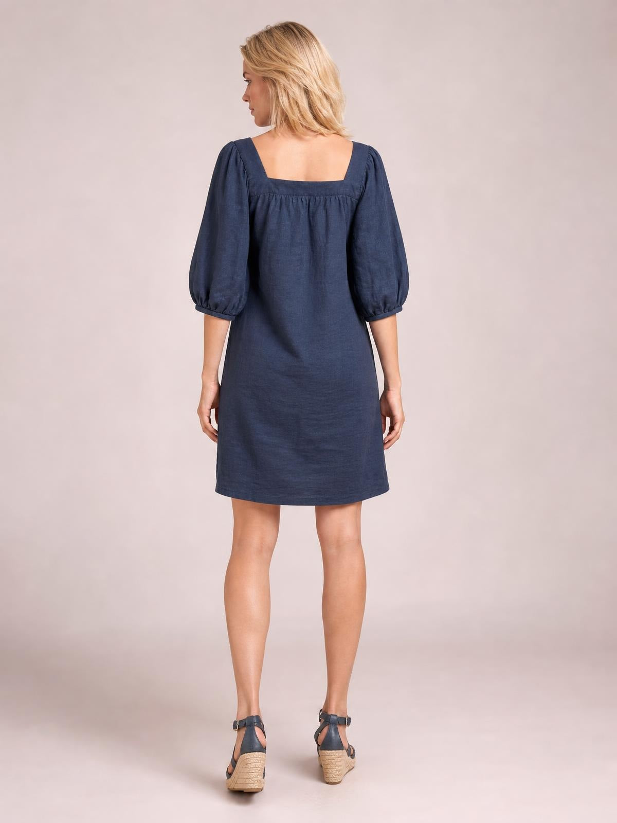 A model stands facing away in a navy square-neck mini dress with voluminous puff sleeves, a relaxed straight silhouette, and gathered seam detail at the back.