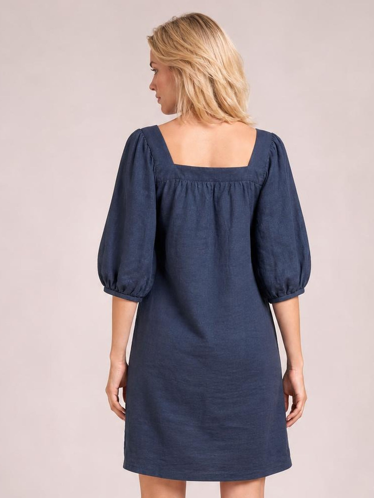 A model stands facing away in a navy square neck mini dress with relaxed silhouette, puff sleeves, gathered seam detail, and a straight hem.