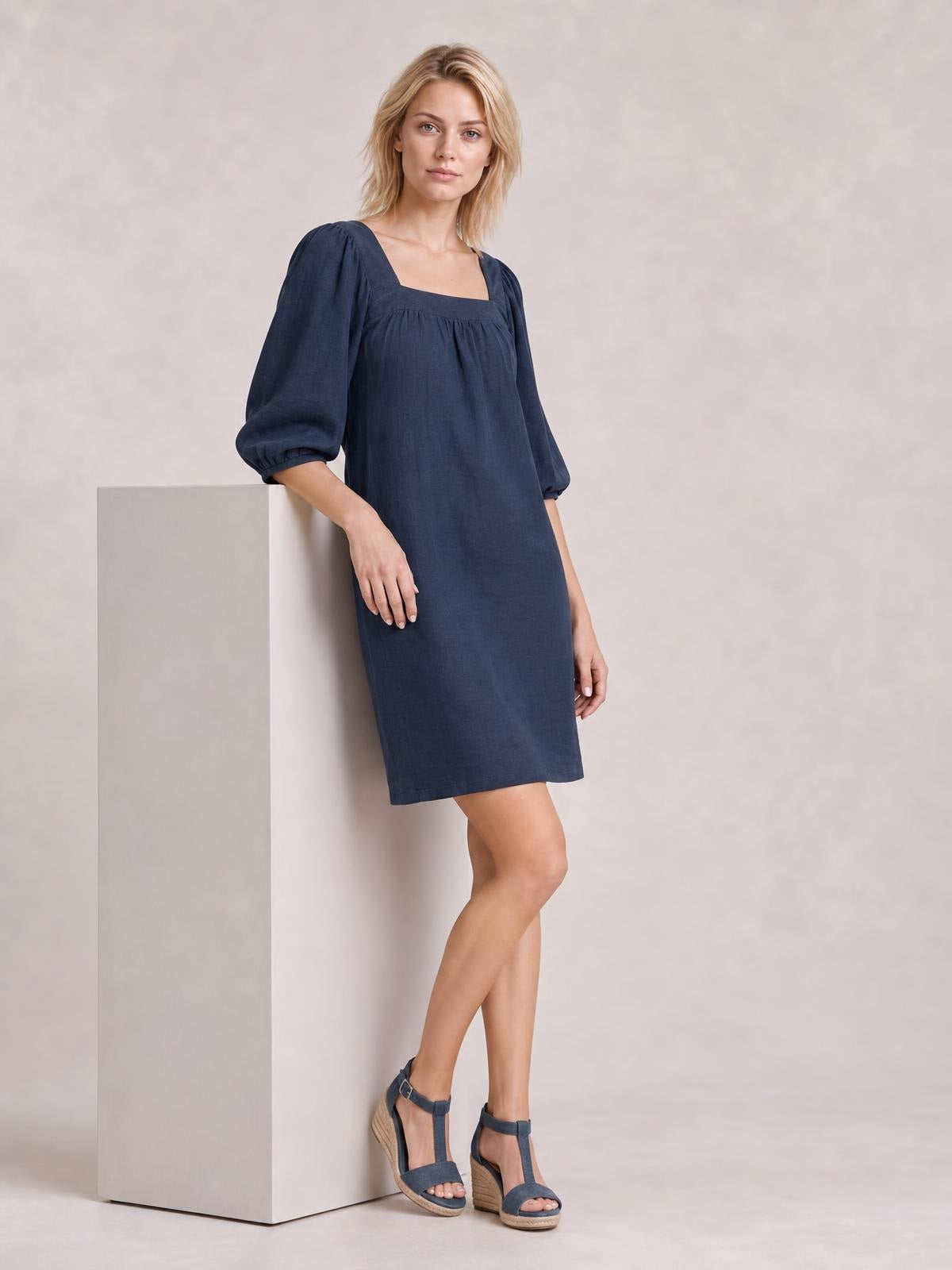 Model in a navy square neck mini dress with puff sleeves, gathered seam detail, and a relaxed straight silhouette, leaning with crossed legs.