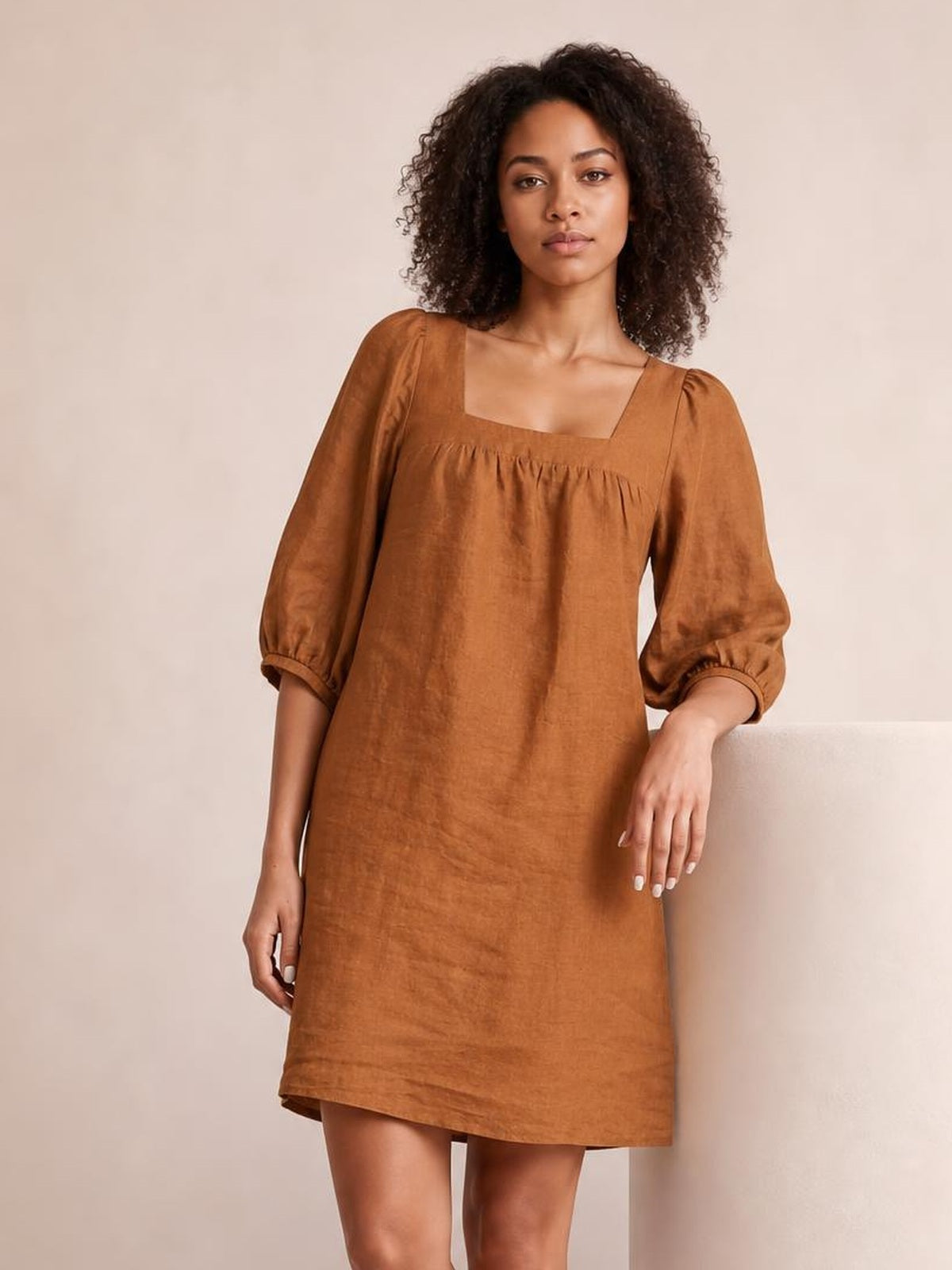 Model in a relaxed pose wearing a rust-brown square neck mini dress with puff sleeves, gathered seam detail, elastic cuffs, and a straight silhouette.