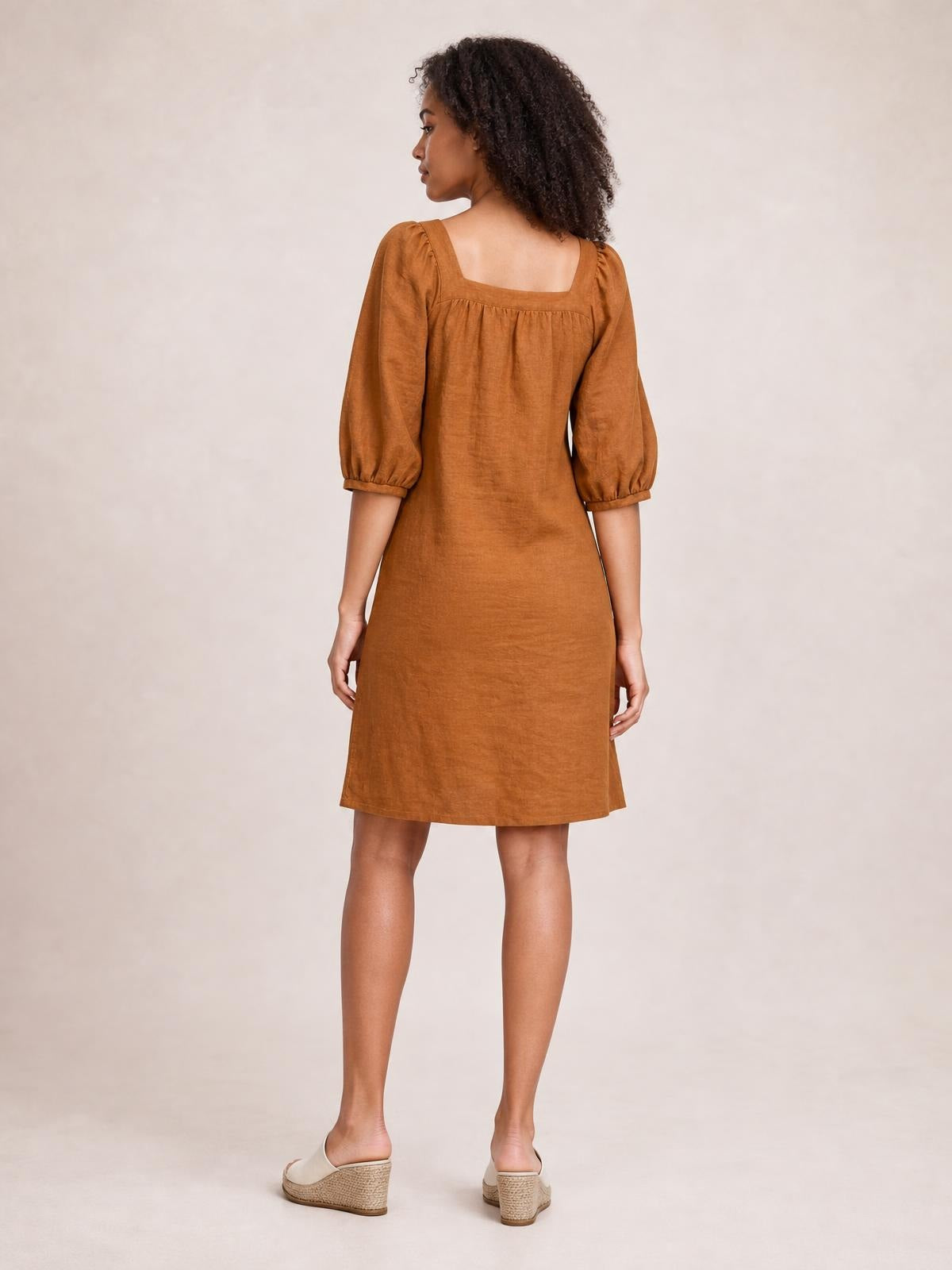 Model viewed from the back in a relaxed rust-brown mini dress with a square neckline, puff sleeves, gathered seam detail, and straight silhouette.