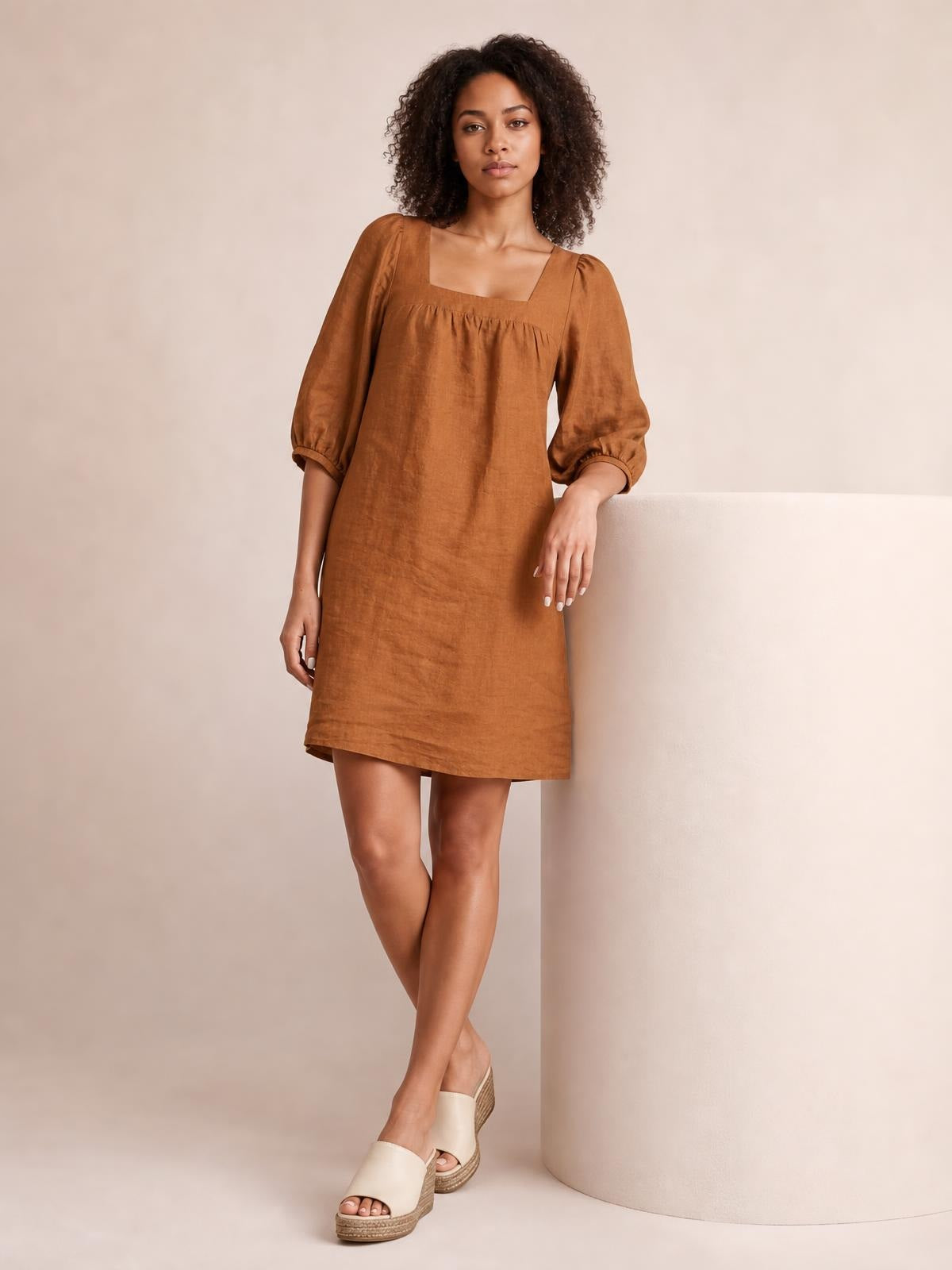 Model wears a rust-brown square-neck mini dress with puff sleeves, seam detailing, and a relaxed straight silhouette, posing with crossed legs.
