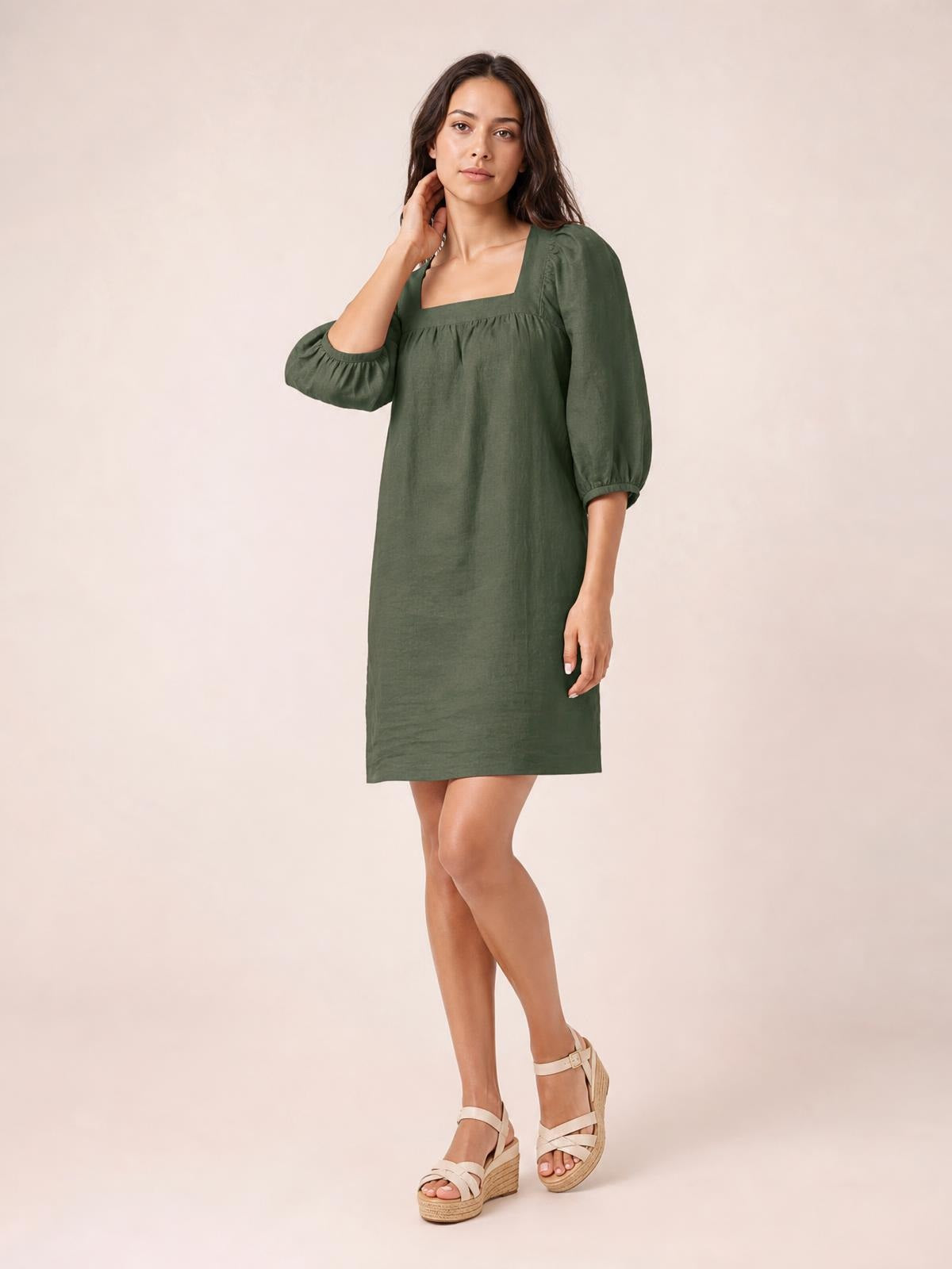 A model poses with one hand at her neck in an olive green square-neck mini dress with puff sleeves, gathered seam detail, and a relaxed straight silhouette.