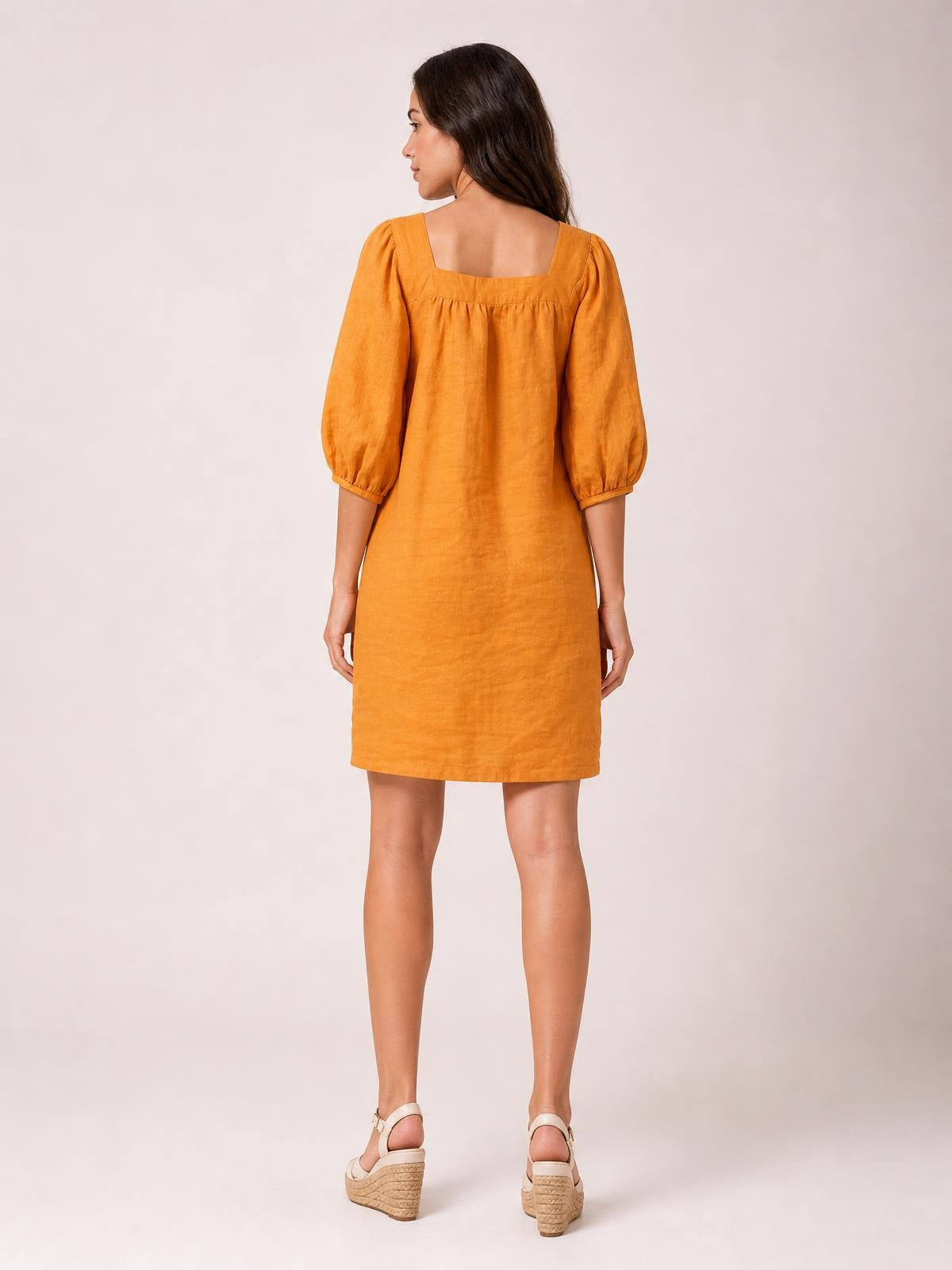 Rear view of a model wearing an orange square-neck mini dress with puff sleeves, seam detailing, and a relaxed straight silhouette.