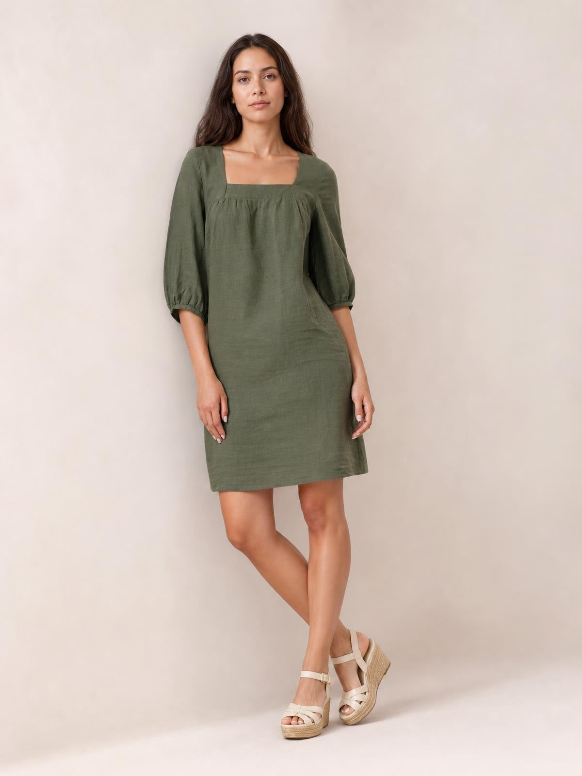 Model wears an olive green square-neck mini dress with gathered seam detail, relaxed silhouette, and three-quarter puff sleeves, posing with crossed legs.