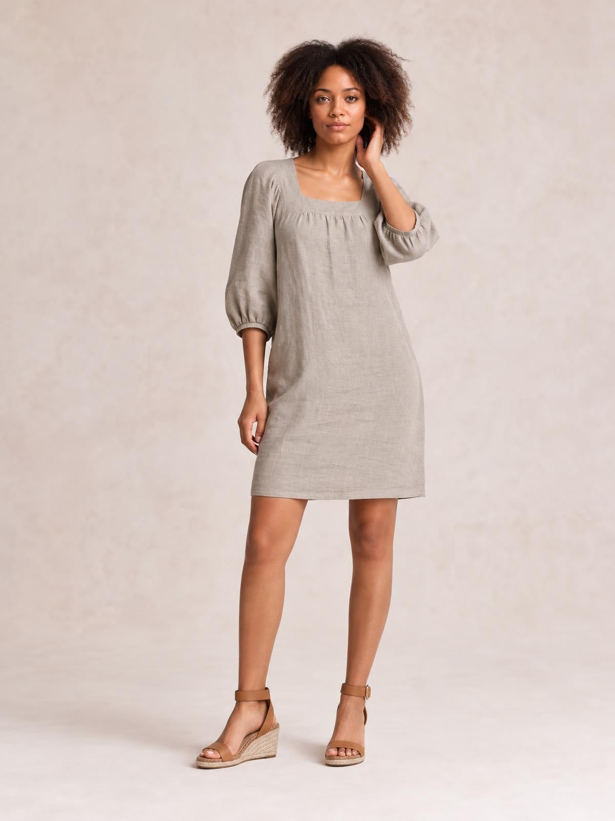 Model wears a taupe square-neck mini dress with gathered puff sleeves, seam detail, and a relaxed straight silhouette, posing with one hand at her hair.
