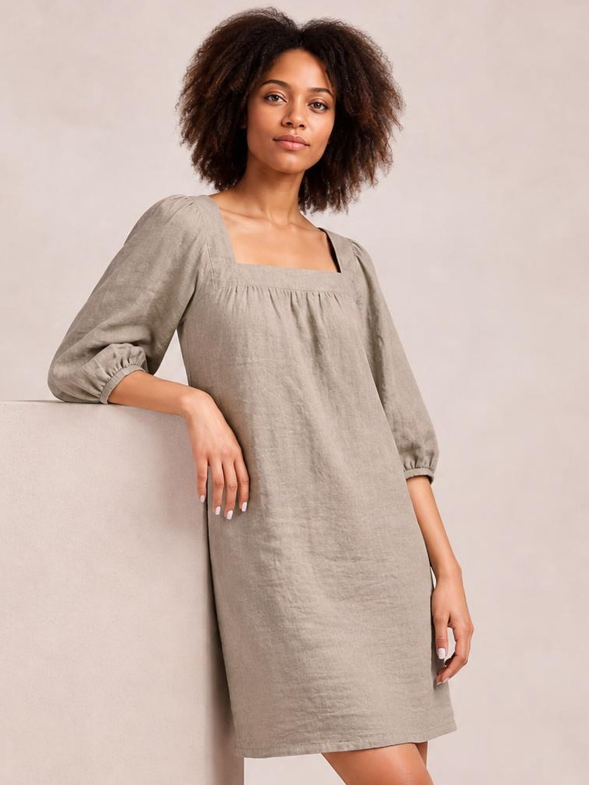 A model leans against a ledge in a taupe square-neck mini dress with puff sleeves, gathered seam detail, and a relaxed straight silhouette.