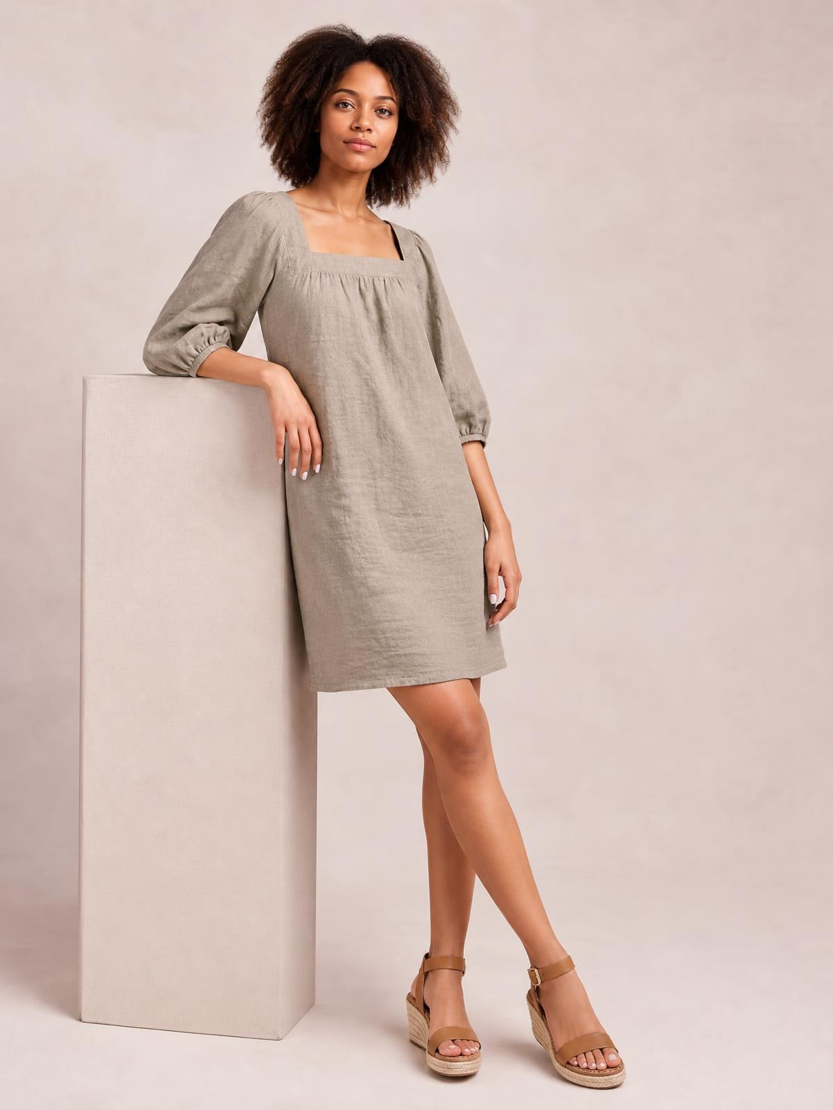 A model wears a taupe square-neck puff sleeve mini dress with gathered cuffs, seam detail, and a relaxed straight silhouette while leaning casually.