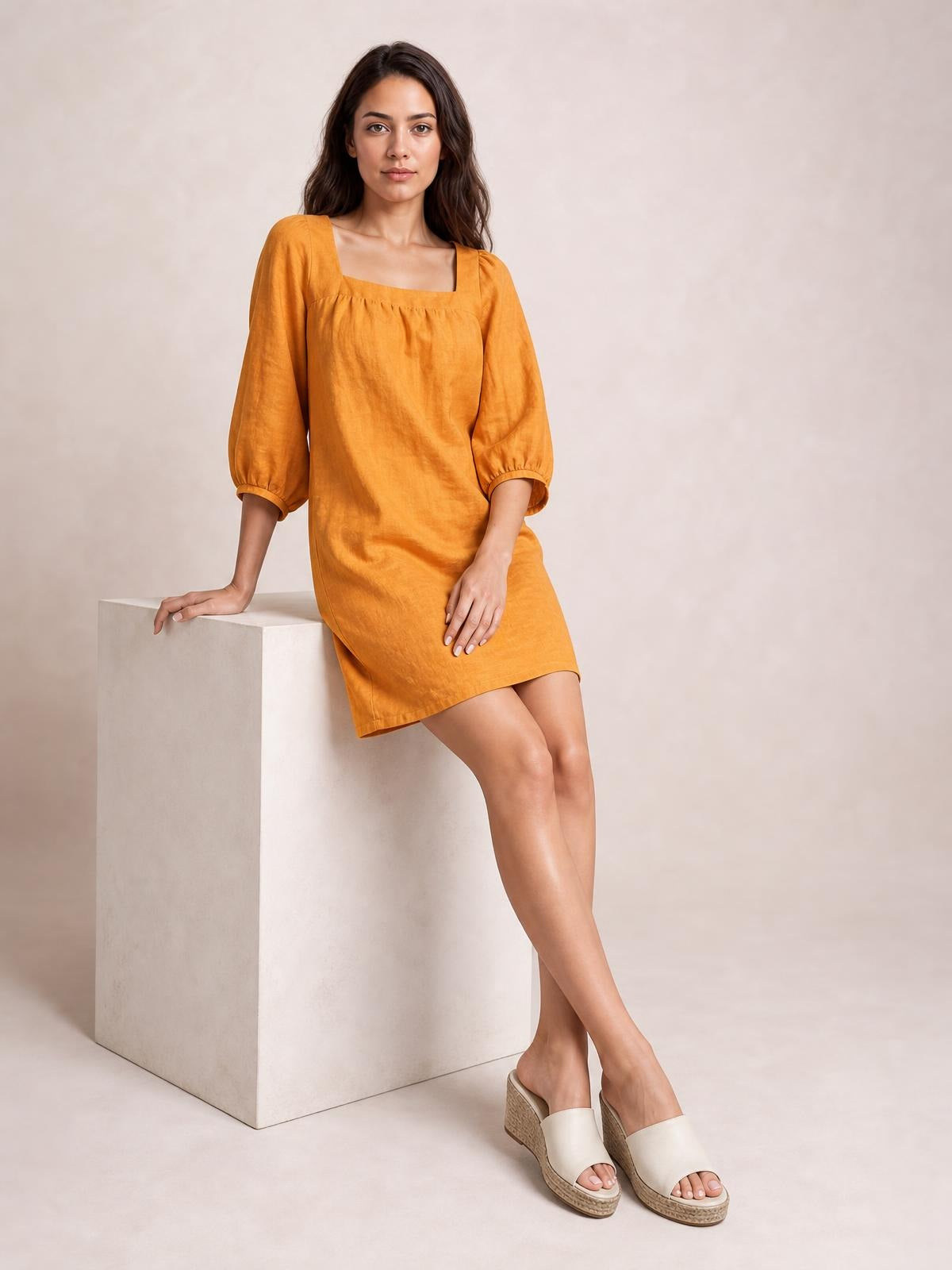 Model seated in a relaxed pose wears a mustard square-neck puff sleeve mini dress with gathered seam detail and a straight, easy silhouette.